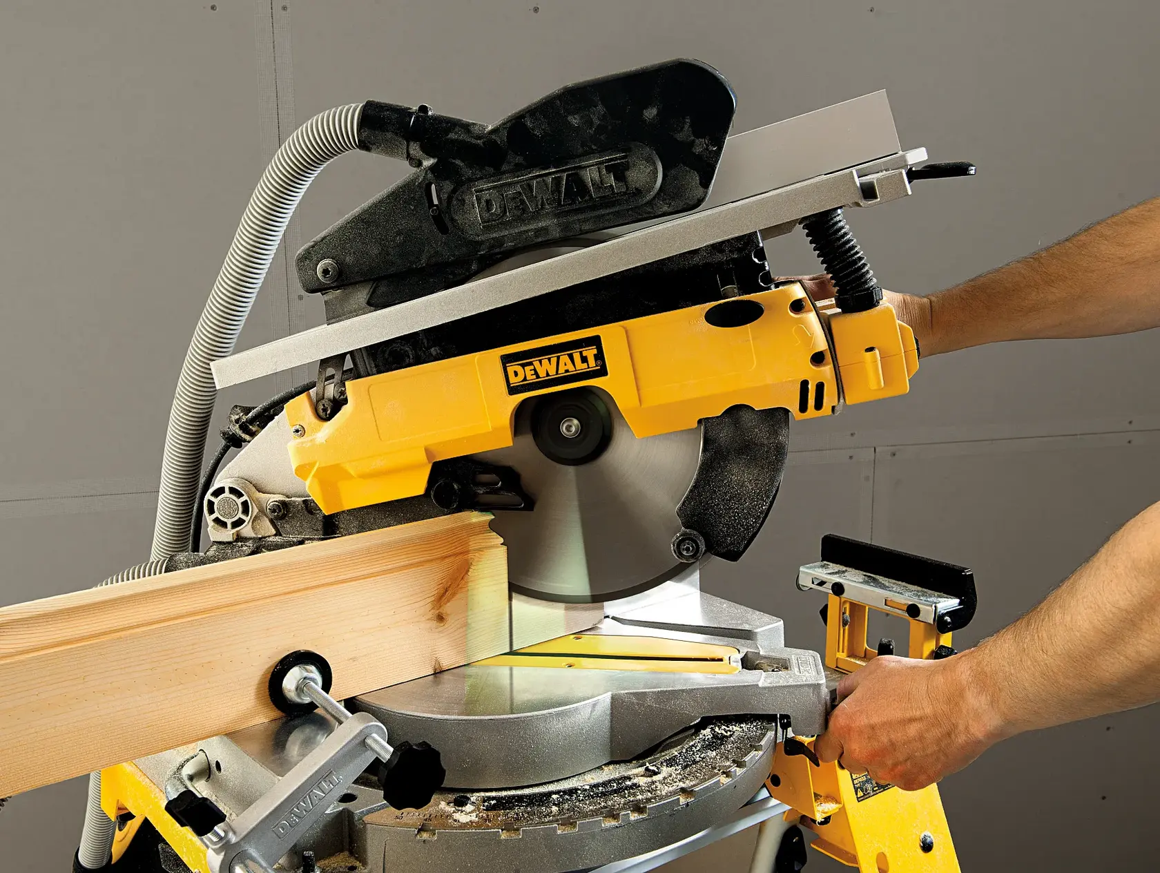 A person is using a DEWALT compound miter saw (model D27113) to cut a piece of wood. The saw is yellow and mounted on a workbench.