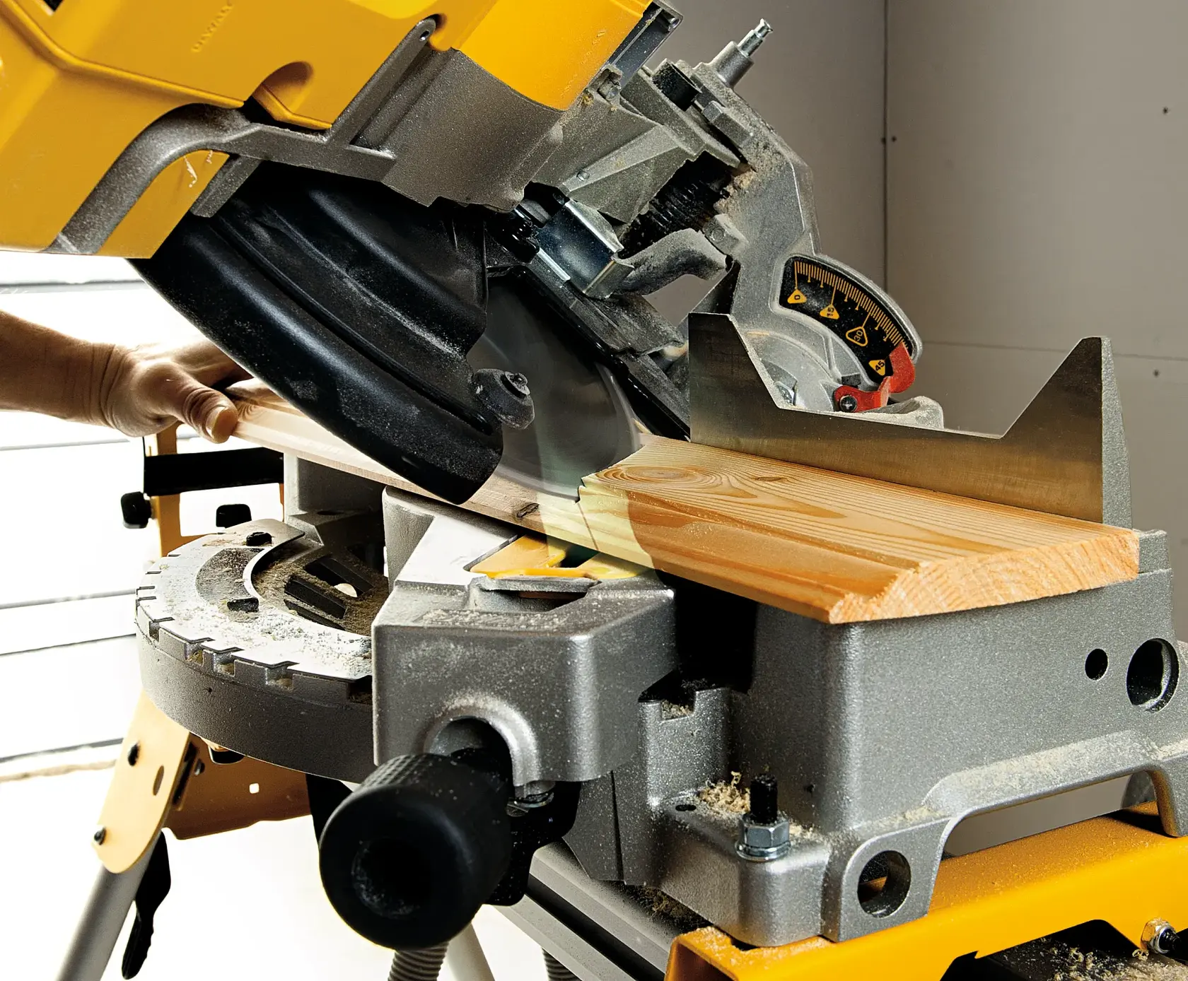 A close-up of a DEWALT miter saw cutting a piece of wood at an angle. The saw is mounted on a workbench, and a person's hand is holding the wood piece in place. Sawdust is visible on the surface.
