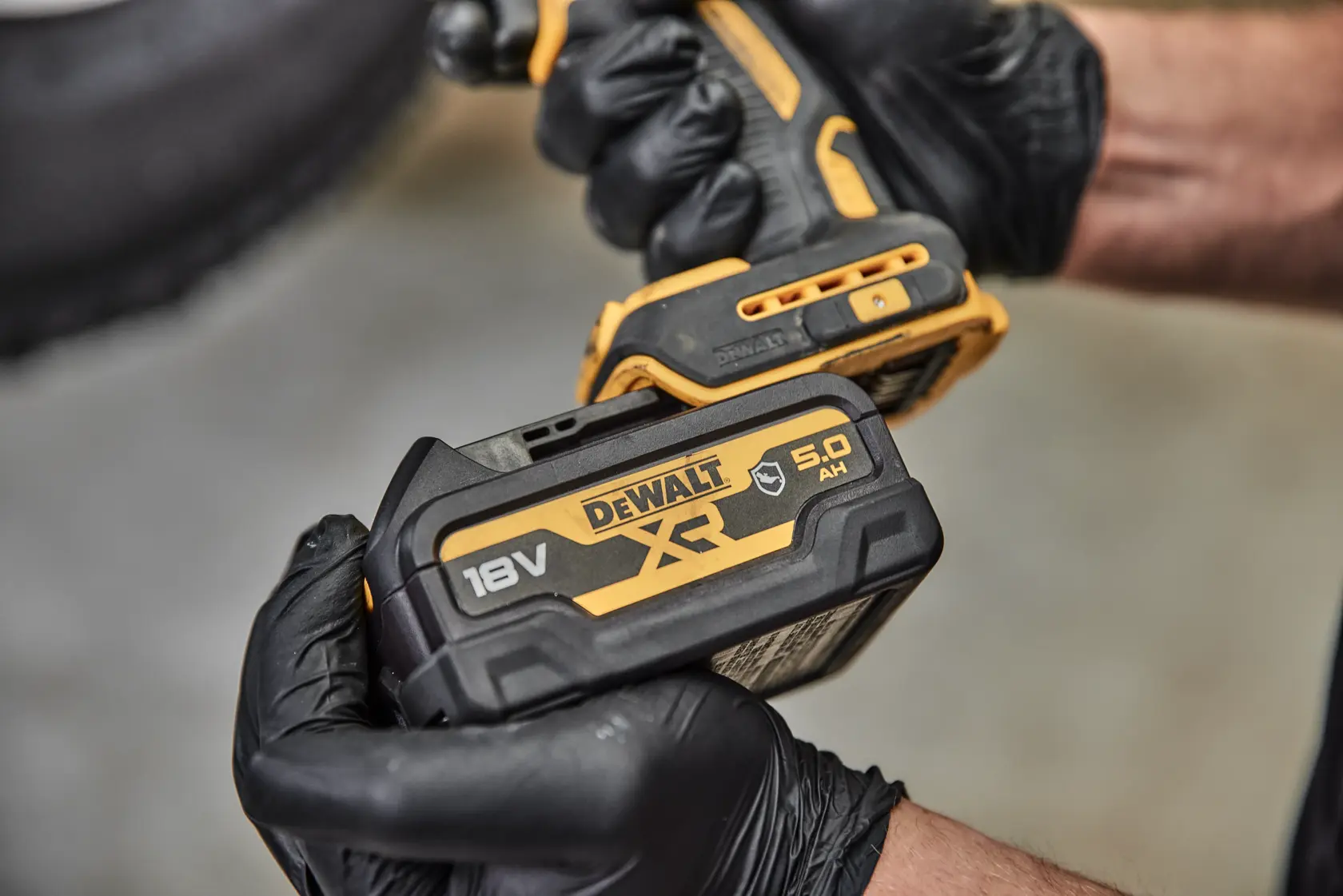 A close-up view of a person wearing black gloves holding a DEWALT cordless power tool with an attached DCB184G 18V XR lithium-ion battery.