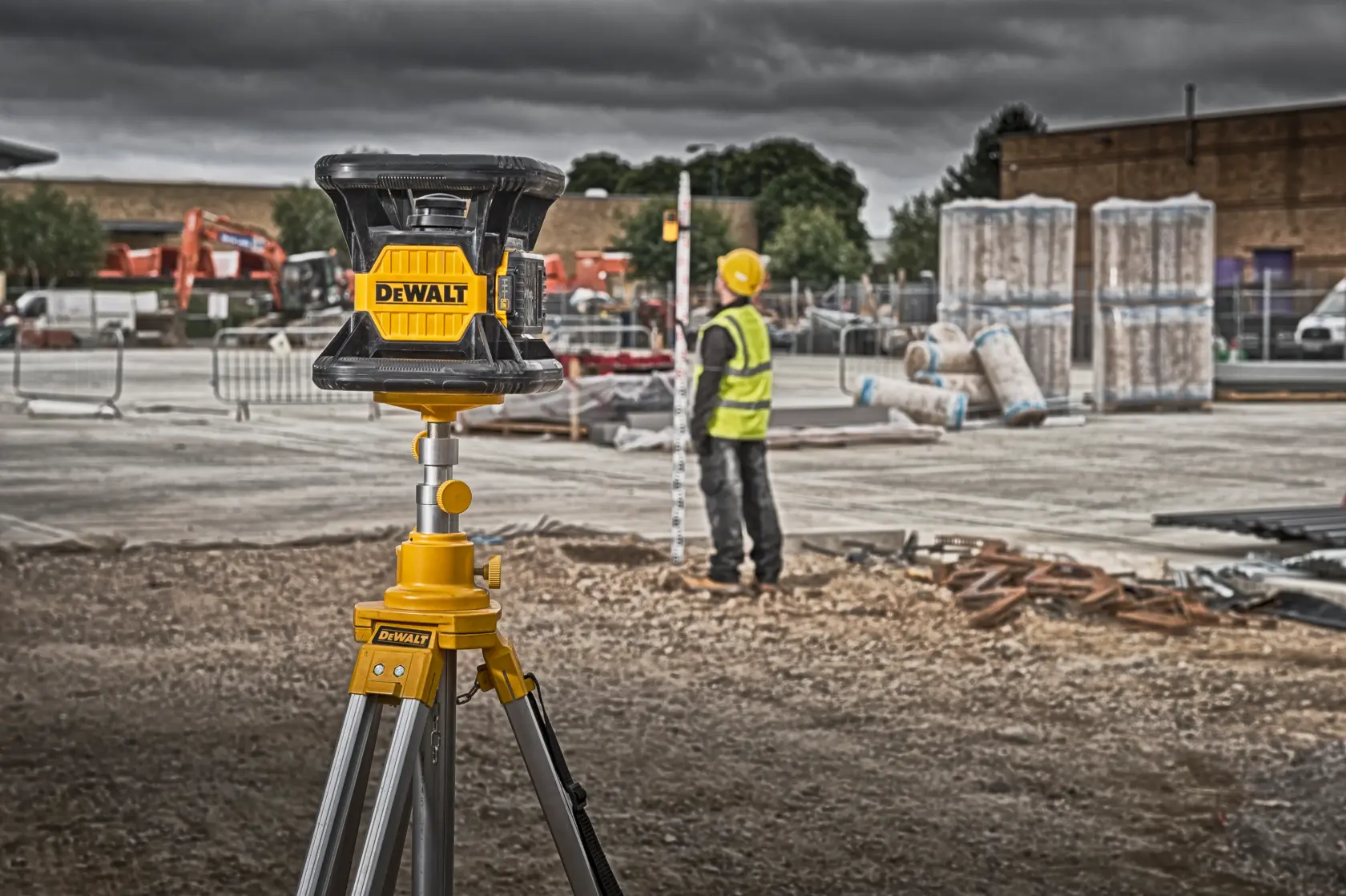 Un nivel láser DEWALT está montado en un trípode en un sitio de construcción. Al fondo, un trabajador con casco amarillo y chaleco reflectante está junto a equipos de medición, rodeado de materiales y maquinaria de construcción.