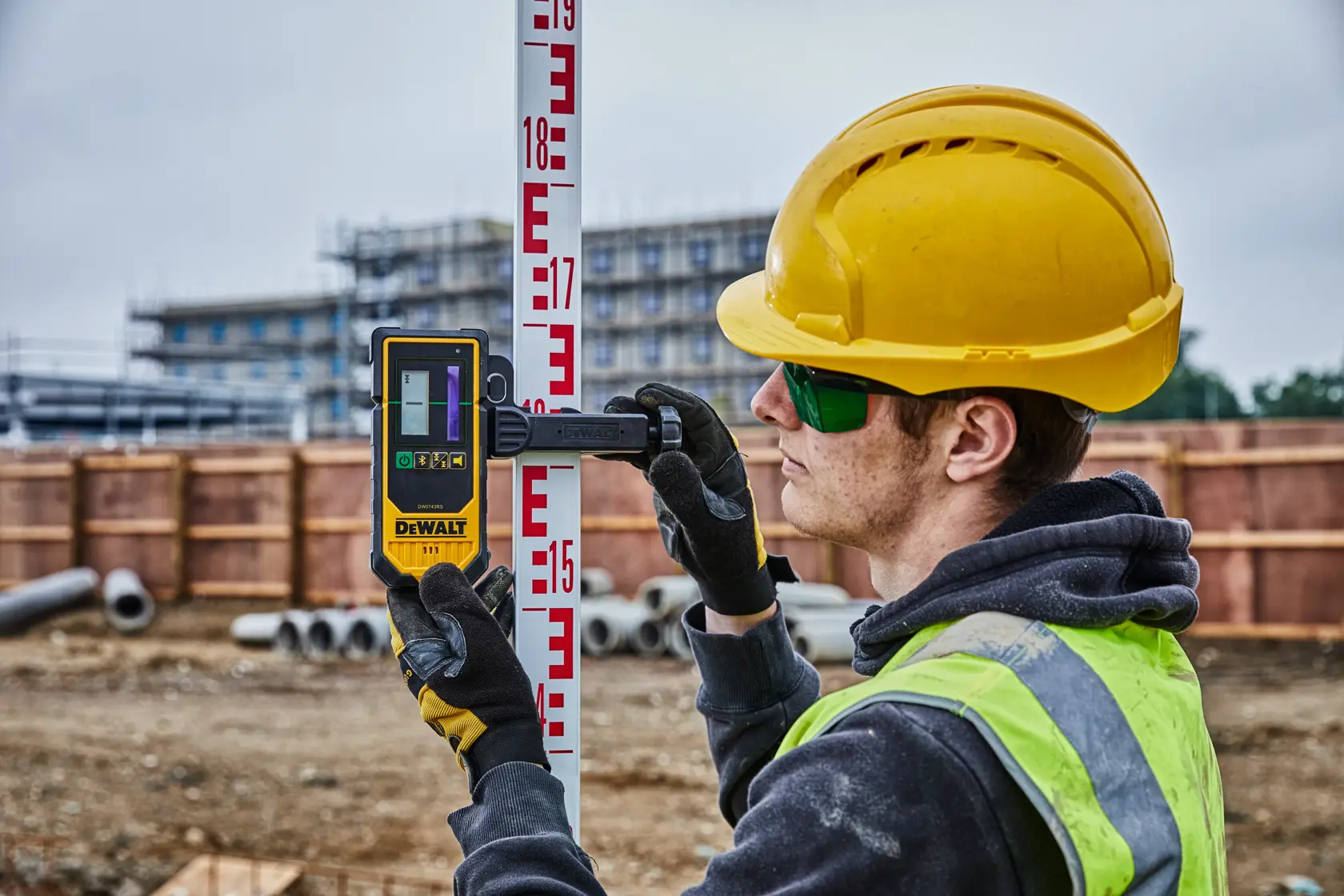 Ein Bauarbeiter mit gelbem Schutzhelm und Handschuhen hält einen DEWALT DCE080D1GS Grade-Rod-Detektor und befestigt ihn an einer Messlatte auf einer Baustelle. Im Hintergrund sind Baumaterialien, Rohre und ein unfertiges Gebäude zu sehen.