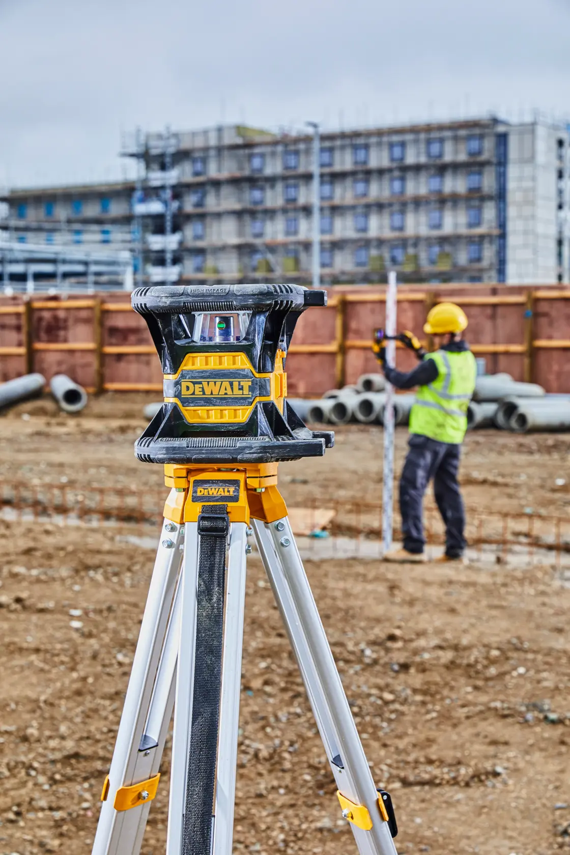 Ein DEWALT-Laser-Nivelliergerät auf einem Stativ auf einer Baustelle, im Hintergrund misst ein Arbeiter mit gelbem Schutzhelm und Warnweste.