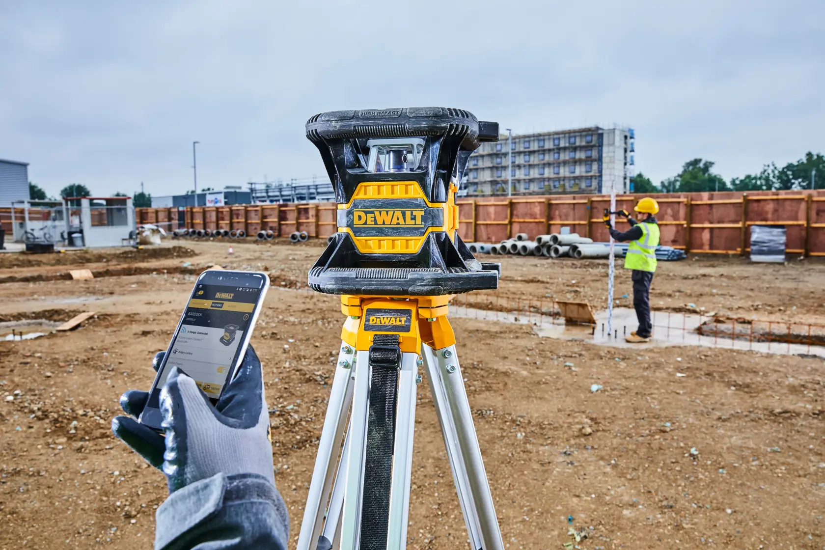 DEWALT-Laser-Nivelliergerät auf einem Stativ auf einer Baustelle, eine behandschuhte Hand hält ein Smartphone und ein Arbeiter mit Sicherheitsweste und Helm misst im Hintergrund.