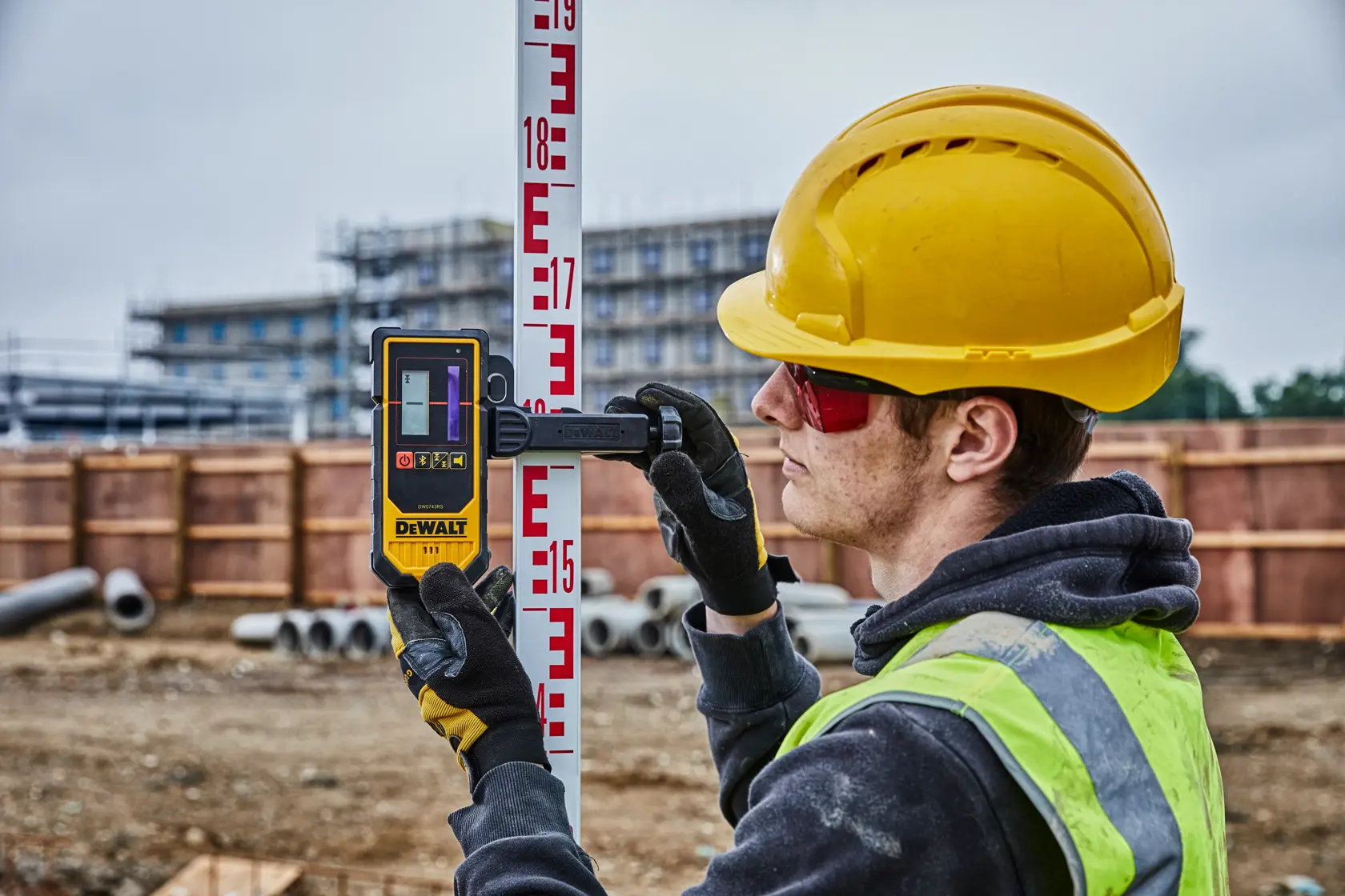 Eine Person auf einer Baustelle verwendet einen DEWALT Laserdetektor, Modell DCE080D1RS, der an einer Nivellierlatte befestigt ist. Die Person trägt einen gelben Schutzhelm und eine Warnweste. Baumaterialien und Bauarbeiten sind im Hintergrund zu sehen.