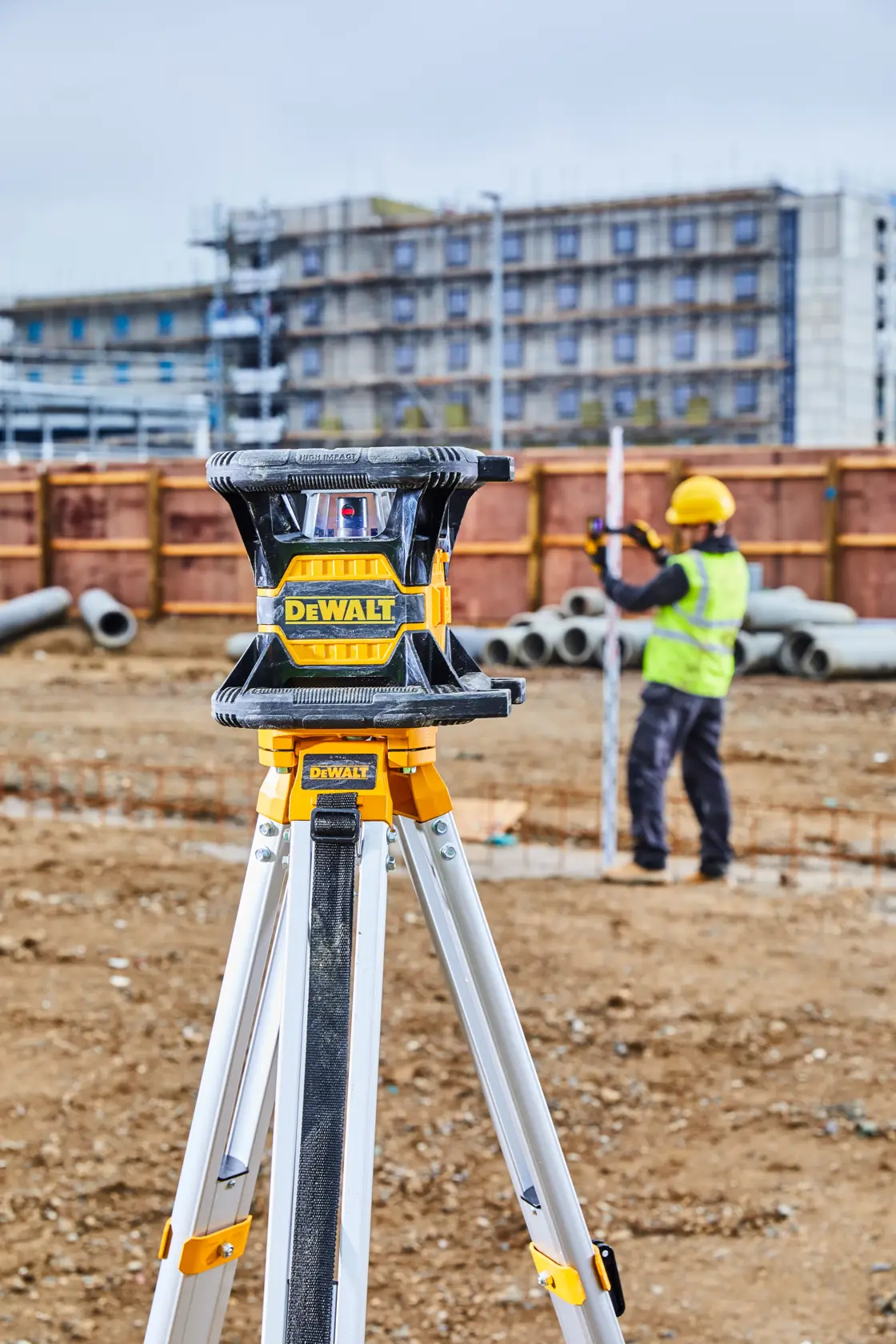 Ein DEWALT-Laser-Nivelliergerät auf einem Stativ steht auf einer Baustelle, während ein Arbeiter mit gelbem Helm und Sicherheitsweste im Hintergrund eine Messlatte einstellt.