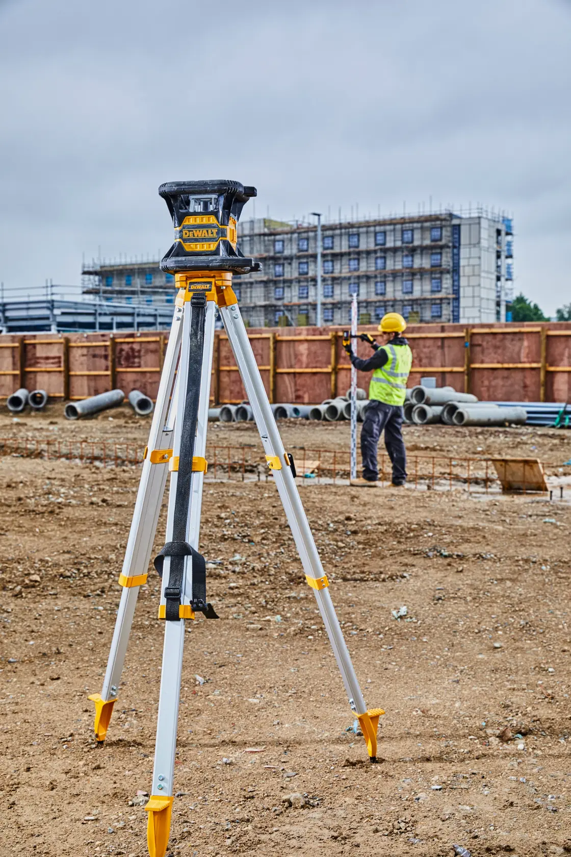 Ein DEWALT-Laser-Nivelliergerät auf einem Stativ steht auf einer Baustelle. Im Hintergrund misst ein Bauarbeiter mit gelbem Helm und Sicherheitsweste mit einer Messlatte, Rohre und ein im Bau befindliches Gebäude sind sichtbar.