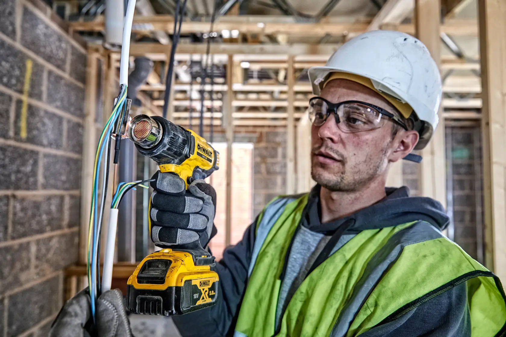 Una persona con equipo de seguridad usa una pistola de calor inalámbrica DEWALT DCE530N para trabajar con cables eléctricos en una obra de construcción. El rostro de la persona está difuminado por motivos de privacidad.
