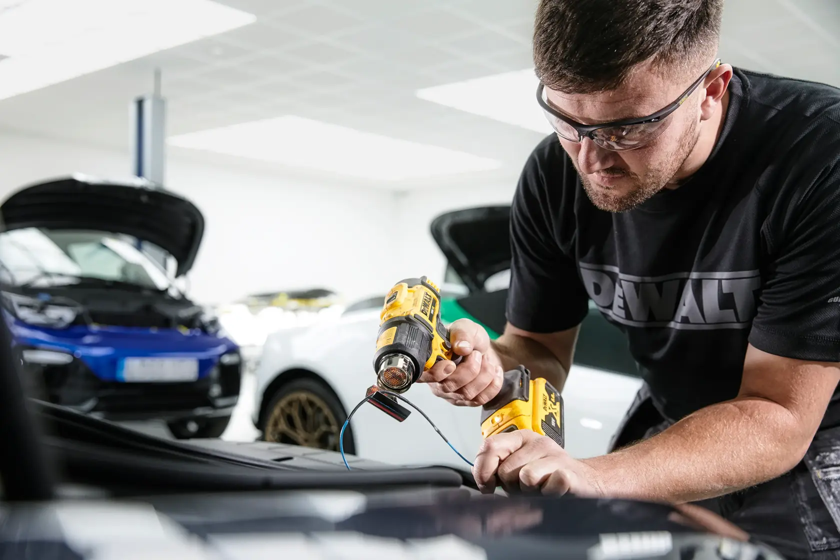 Una persona utiliza una pistola de calor inalámbrica DEWALT (modelo DCE530N) en una pieza de coche, con el rostro difuminado por privacidad. El individuo lleva una camiseta DEWALT y trabaja en un garaje con coches de fondo.