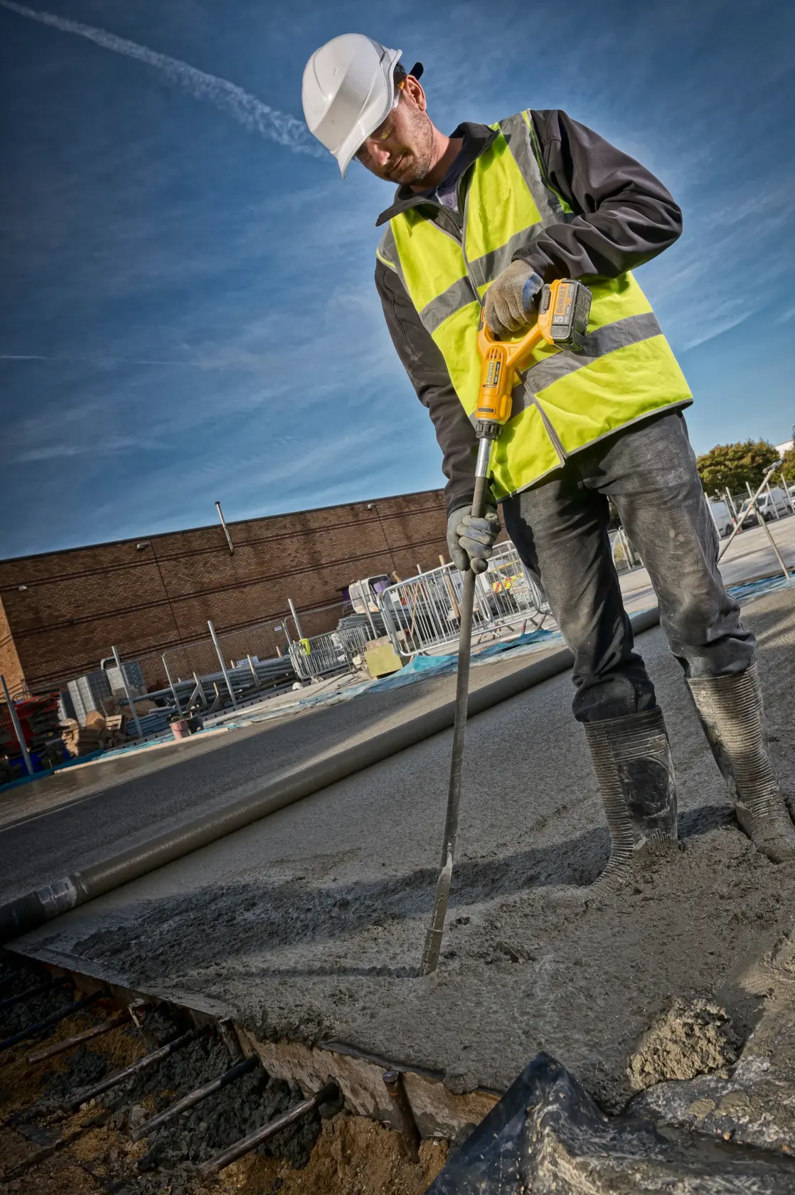 Ein Arbeiter mit Schutzhelm, Warnweste, Handschuhen und Stiefeln bedient ein DEWALT DCE531N Werkzeug, um frisch gegossenen Beton auf einer Baustelle zu verdichten.