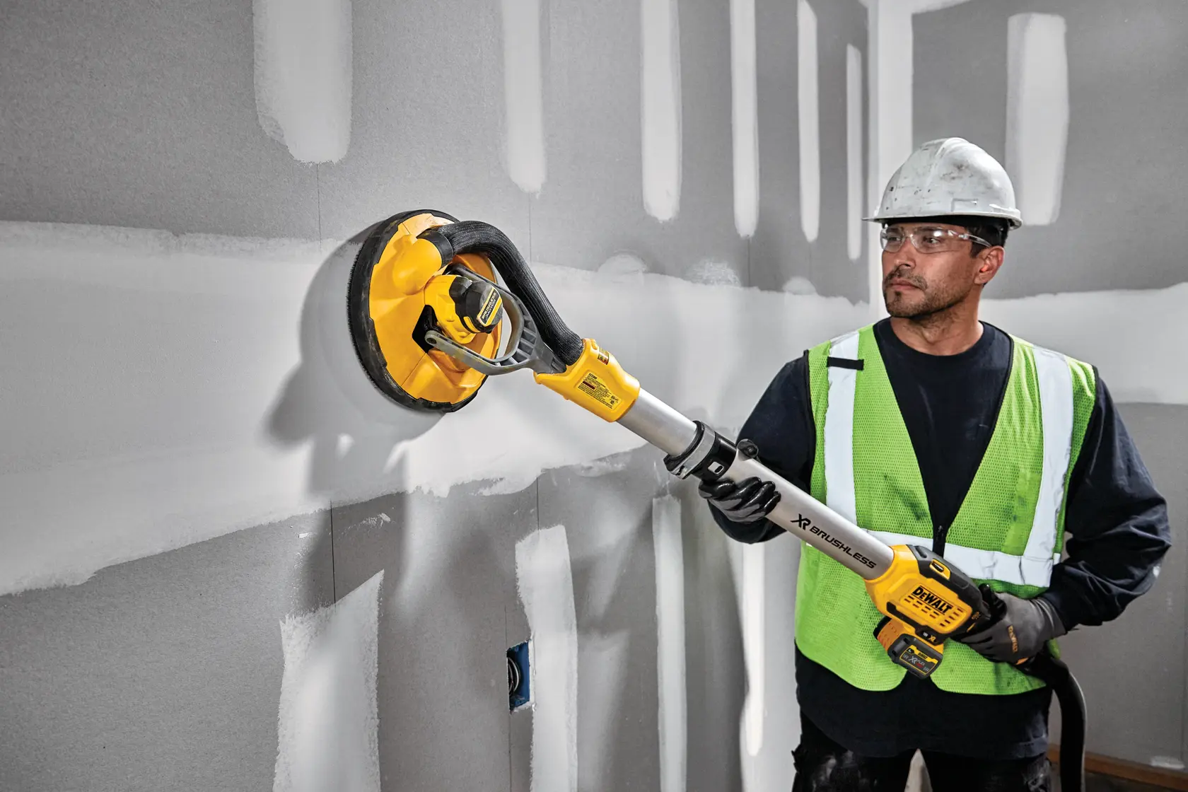 Una persona con casco y chaleco de seguridad está usando una lijadora para paredes de yeso inalámbrica DEWALT DCE800T2 sobre una pared con juntas de yeso sin terminar.