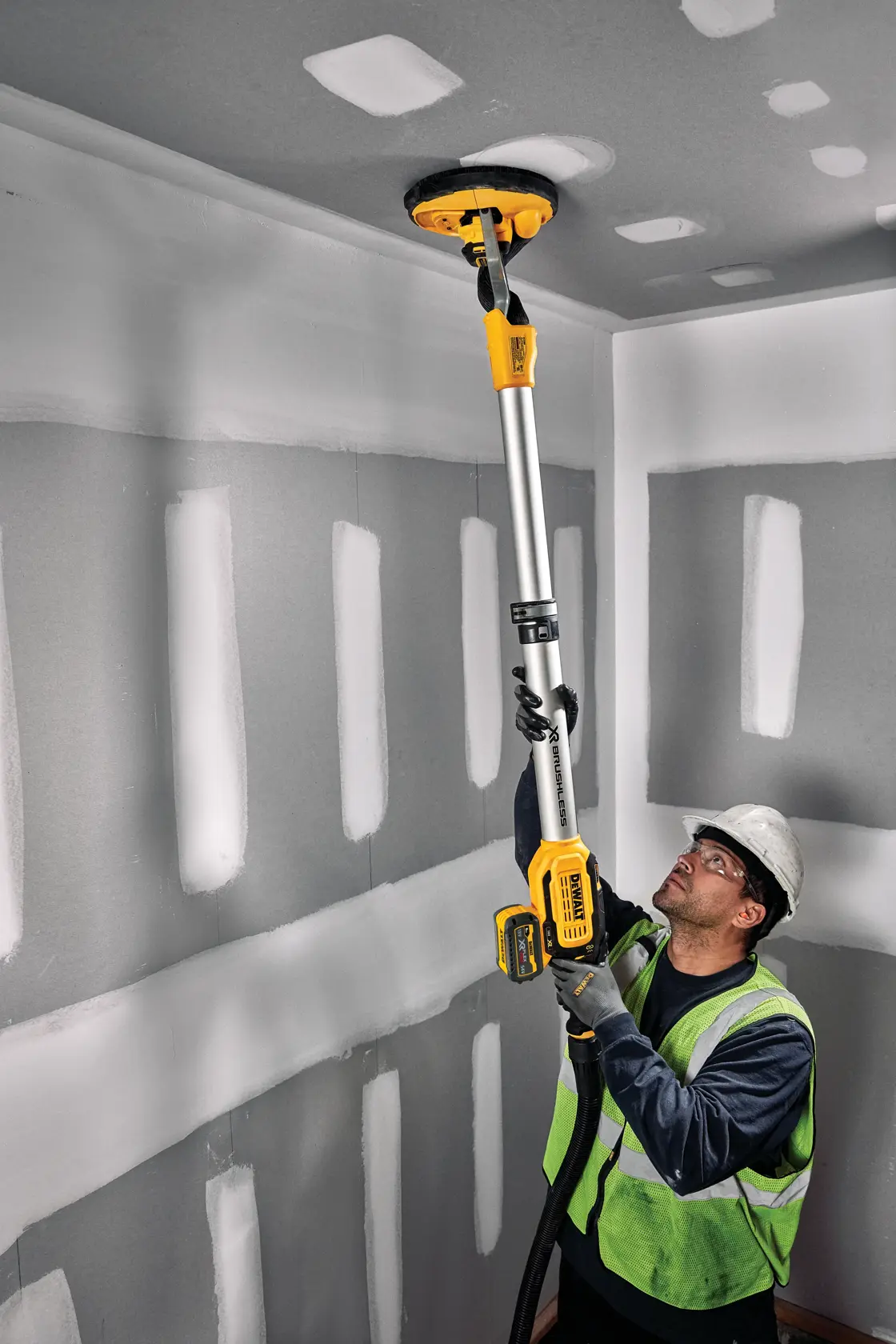 Un trabajador con chaleco de seguridad y casco usa una lijadora inalámbrica para paneles de yeso DEWALT con un mango extensible para lijar el techo en una habitación con paneles de yeso sin terminar.