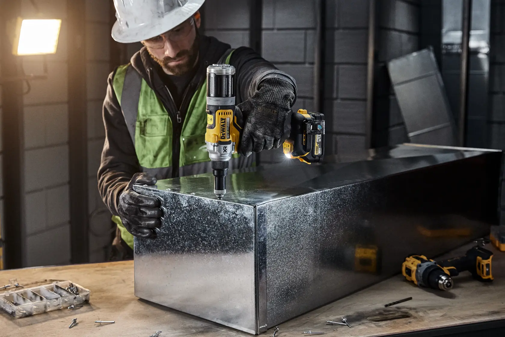 A person wearing protective gear uses a DEWALT DCF414NT cordless power tool to work on a large metal box in an industrial setting. Various tools and screws are visible on the worktable.