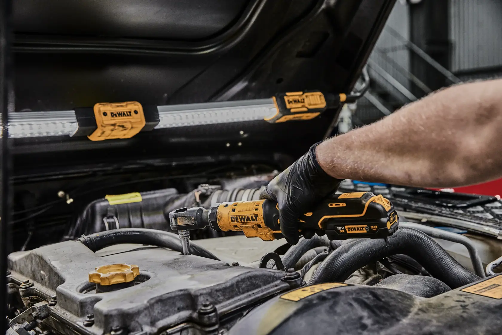 Eine Person mit schwarzen Handschuhen verwendet ein kabelloses DEWALT Ratschenwerkzeug am Motor eines Autos, während eine DEWALT Arbeitsleuchte am geöffneten Fahrzeugmotor befestigt ist.