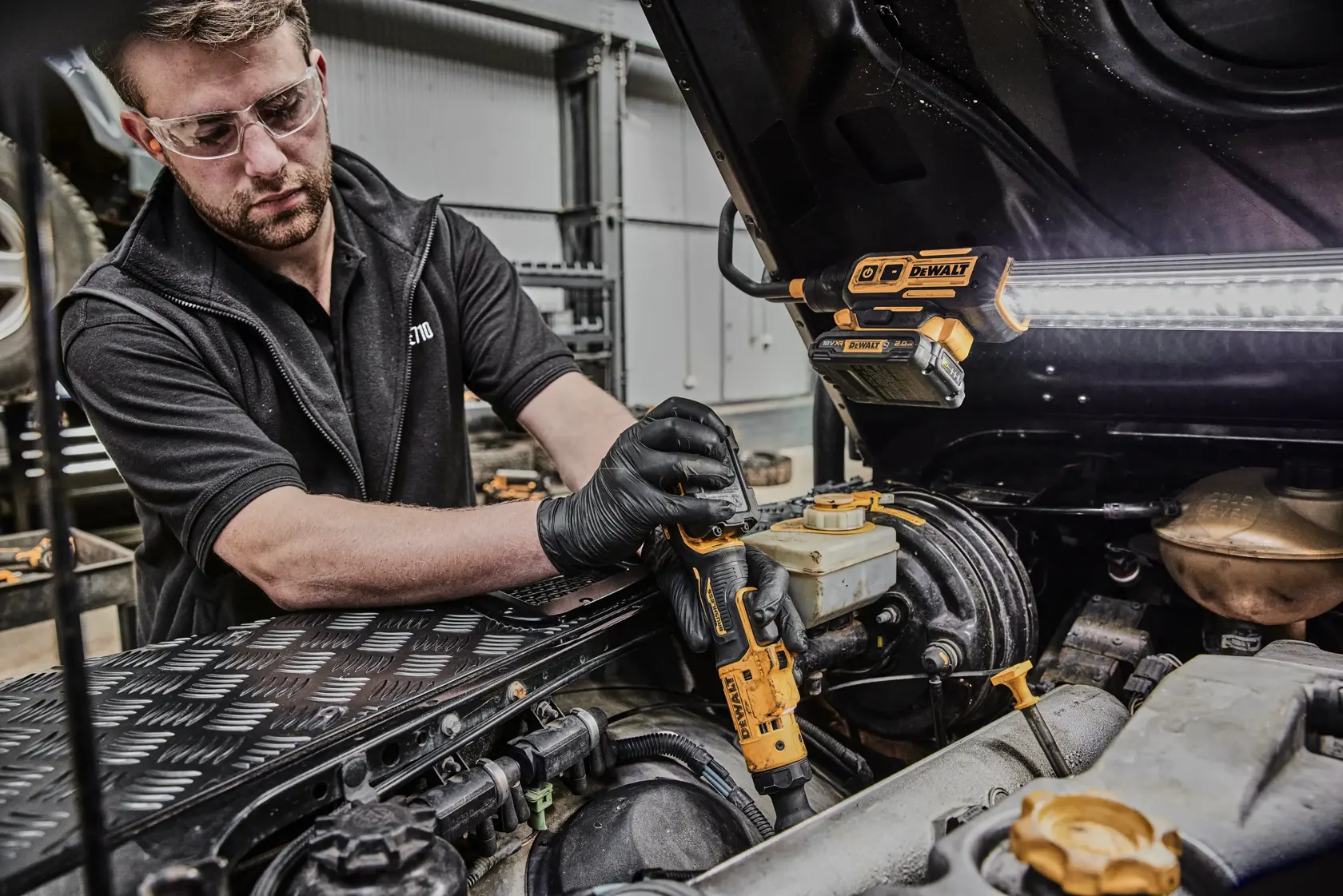 Eine Person mit schwarzen Handschuhen benutzt einen kabellosen DEWALT DCF503N Ratschenschrauber, um an einem Auto-Motor in einer Werkstatt zu arbeiten. Eine DEWALT Arbeitsleuchte ist an der Motorhaube befestigt und beleuchtet den Arbeitsbereich.