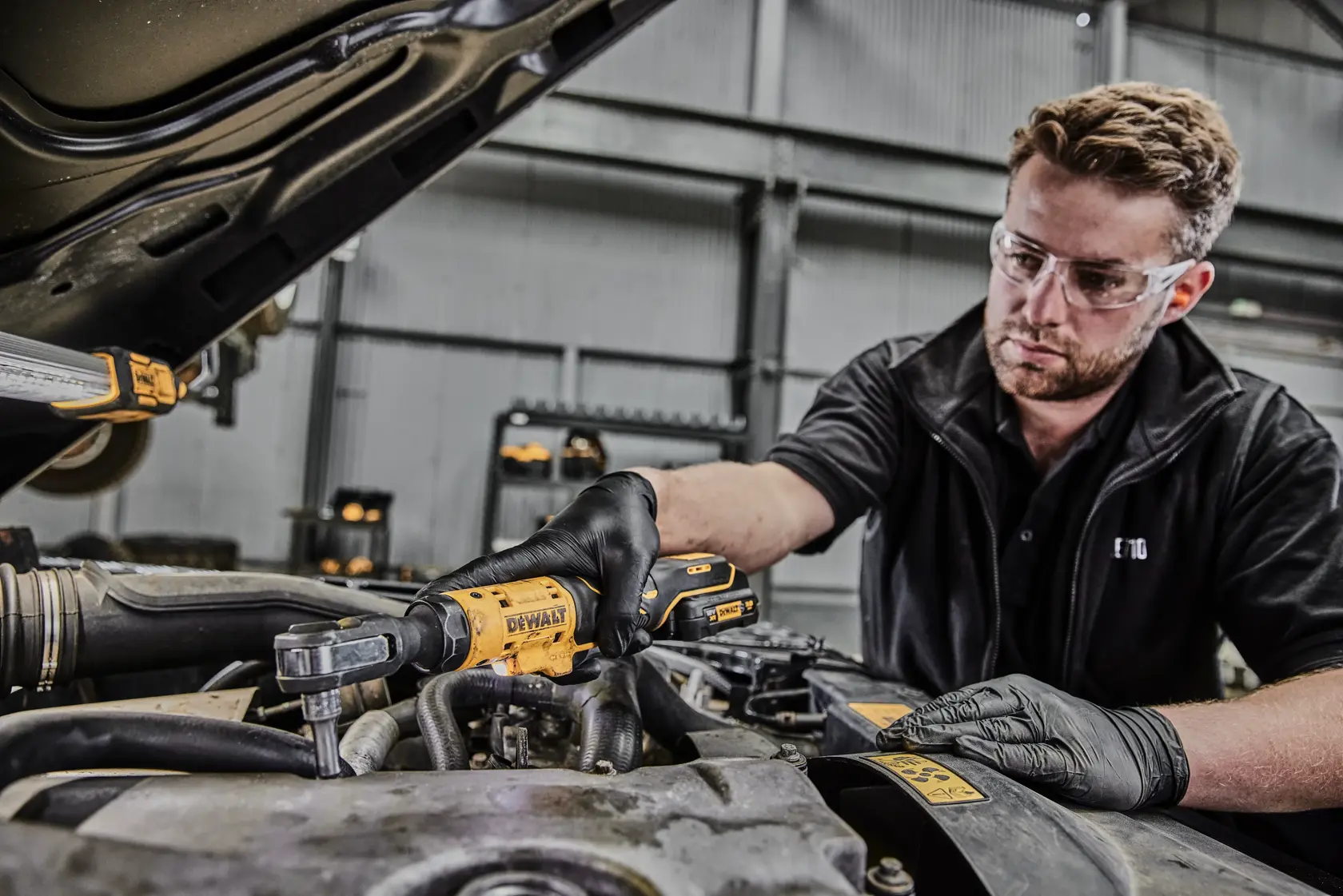 Una persona con guantes negros está utilizando una herramienta de carraca inalámbrica DEWALT DCF504N para trabajar en el motor de un automóvil en un taller industrial. El rostro de la persona está difuminado por privacidad.