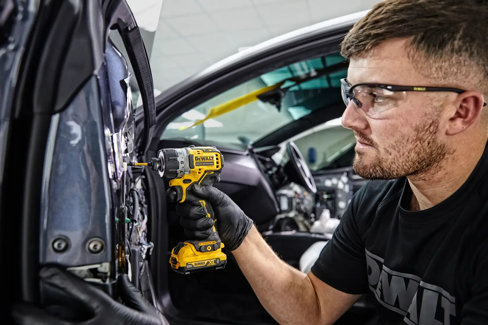 Una persona con guantes negros utiliza un destornillador inalámbrico amarillo DEWALT (DCF601D2) para trabajar en el interior de la puerta de un coche. El rostro de la persona está difuminado y lleva una camiseta DEWALT.