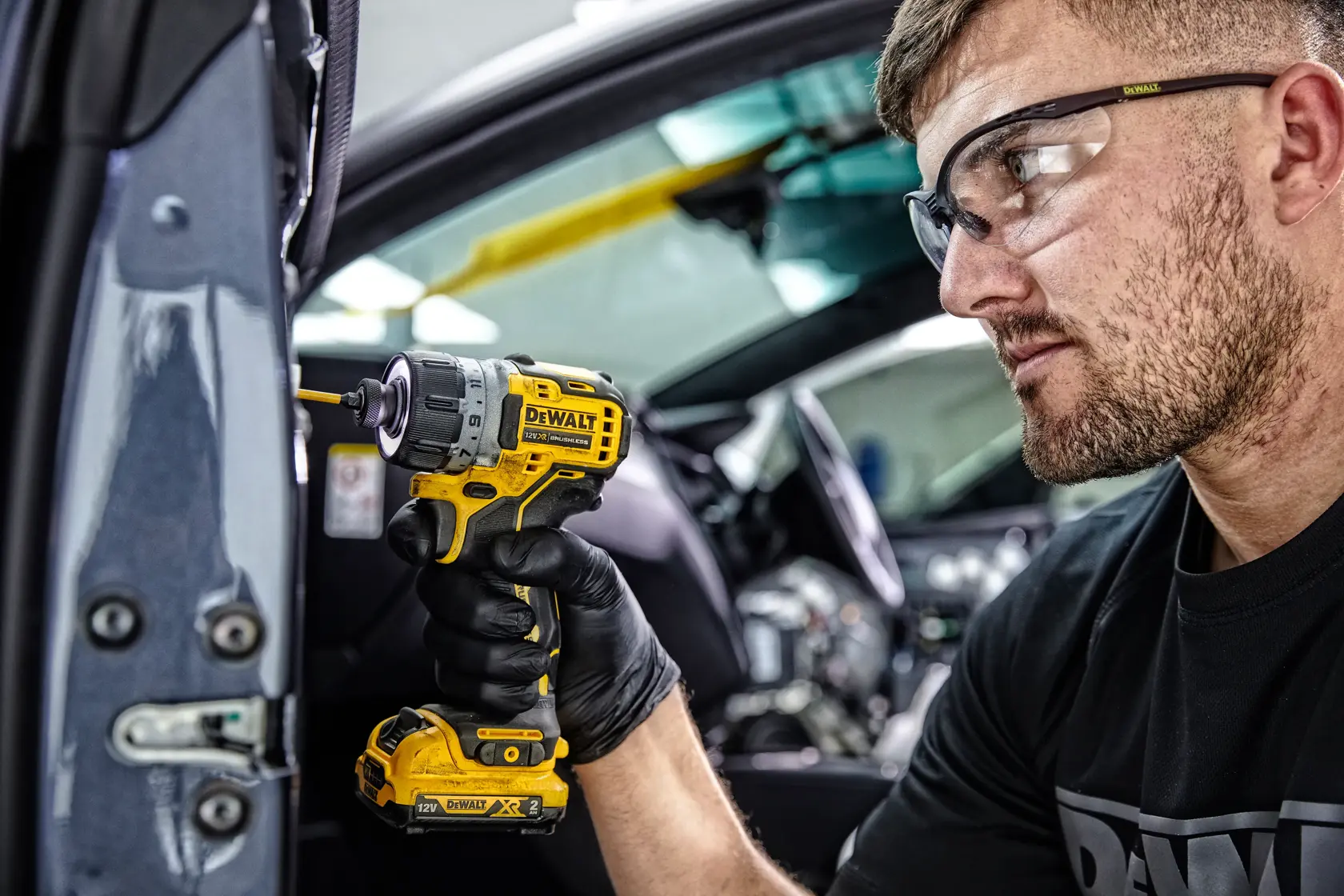 Una persona con guantes negros utiliza un destornillador inalámbrico amarillo DEWALT DCF601D2 para trabajar en una parte dentro de un vehículo. El rostro de la persona está desenfocado por privacidad.