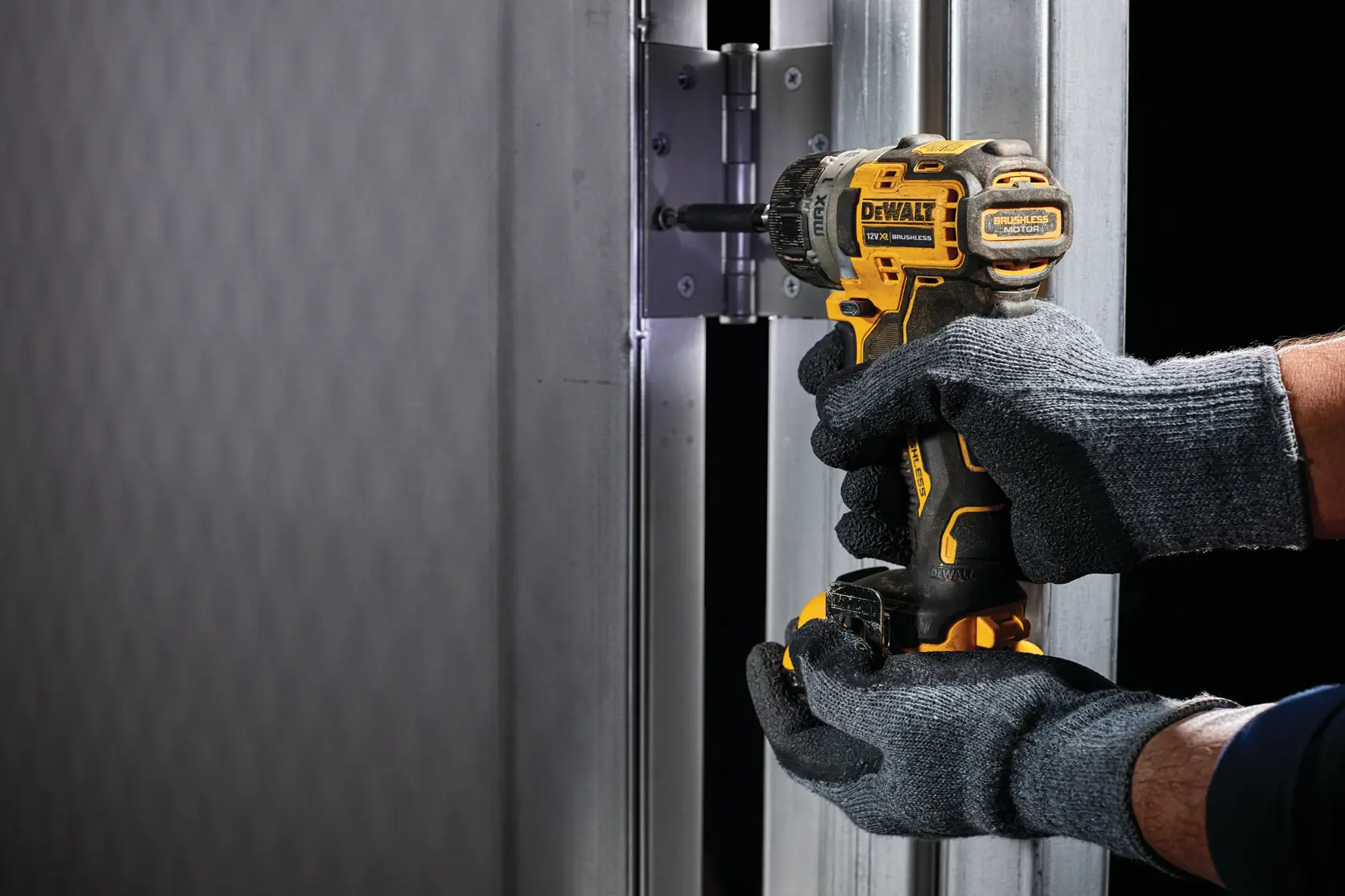 Manos con guantes usan un taladro DEWALT para colocar una bisagra en un marco de puerta metálico.