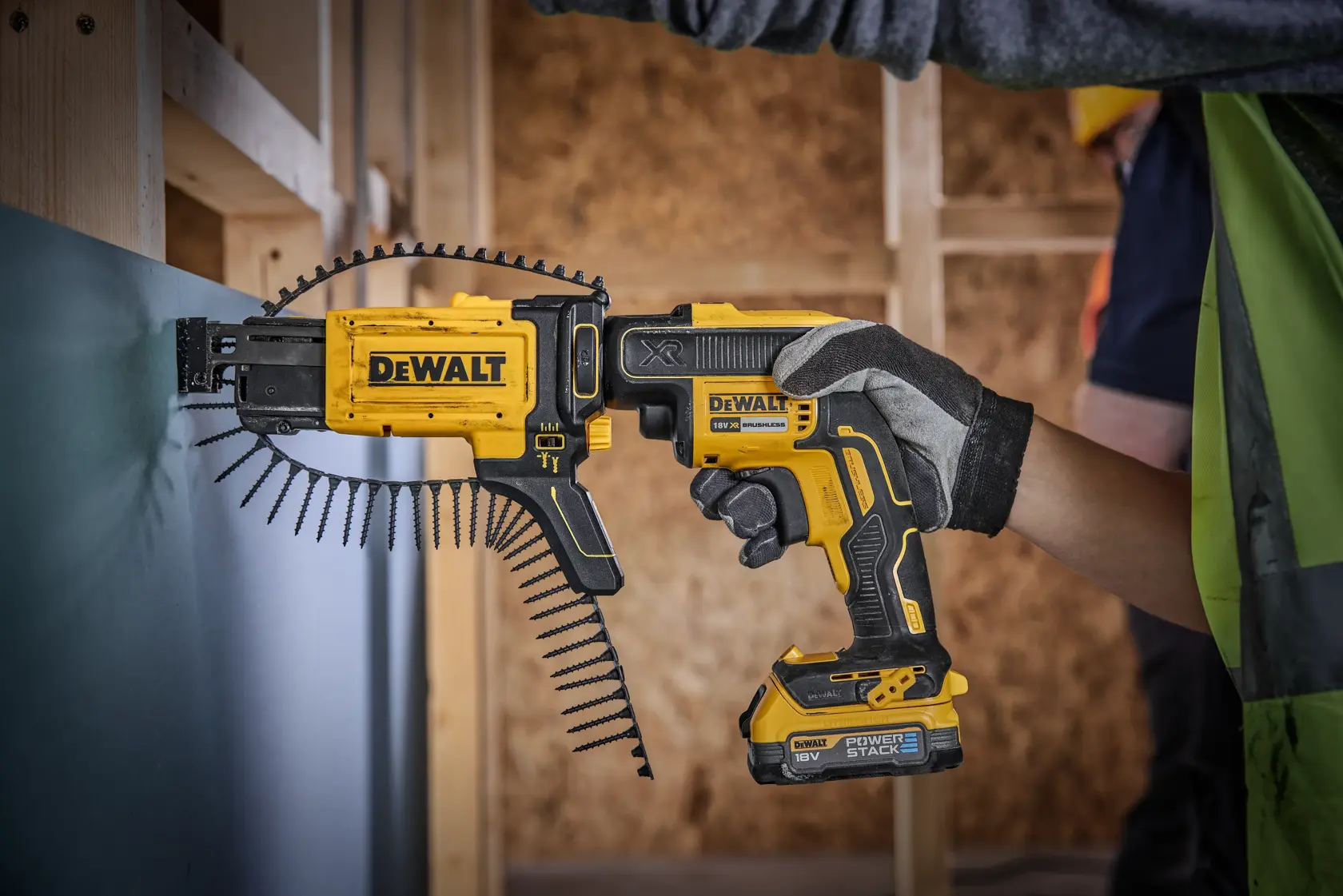 A person wearing gloves uses a DEWALT cordless screw gun with an auto-feed attachment to fasten screws into a wall panel during construction.