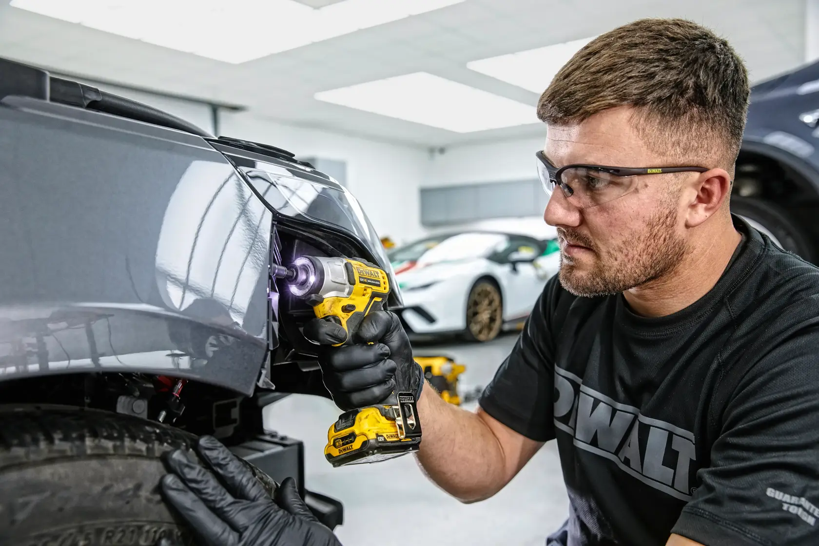 Una persona con guantes negros utiliza un taladro inalámbrico amarillo DEWALT para trabajar en la parte delantera de un automóvil gris en un taller. El rostro de la persona está difuminado por privacidad y hay otros autos al fondo.