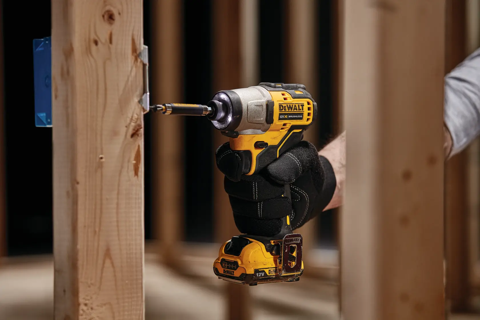 Una persona con guantes negros está usando un destornillador de impacto inalámbrico DEWALT para atornillar en un marco de madera en un entorno de construcción.