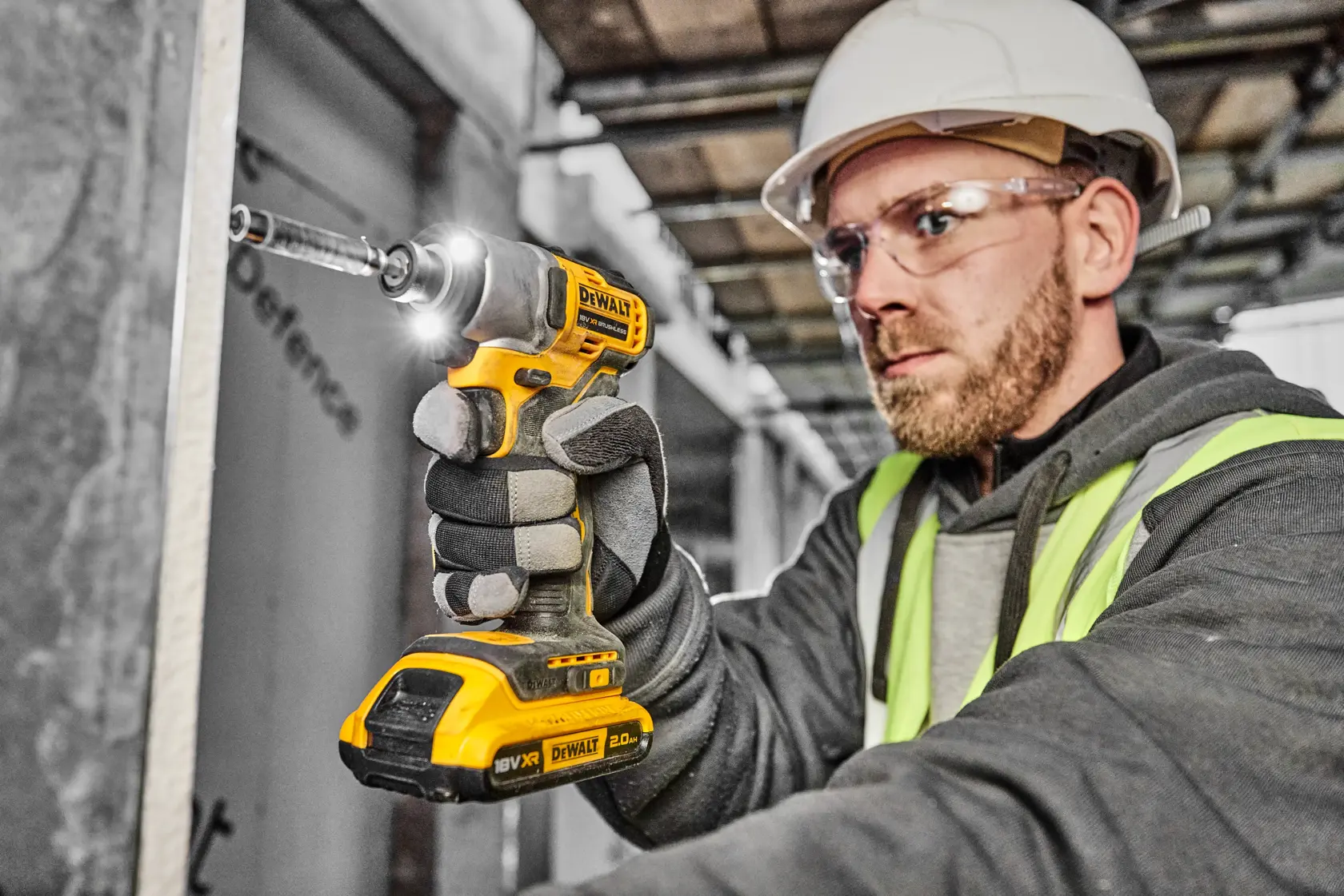 Person wearing a safety helmet and high-visibility vest using a DEWALT DCF840D2T cordless impact driver on a construction site. The person's face is blurred for privacy.
