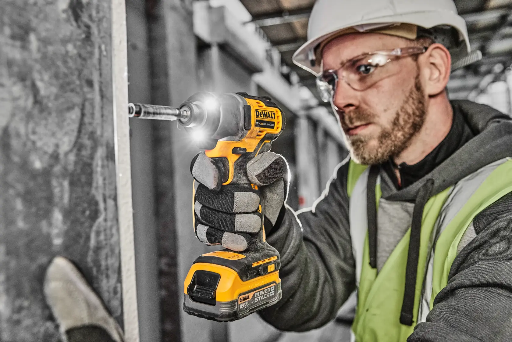 A person wearing work gloves, a safety helmet, and a high-visibility vest is using a DEWALT DCF840 cordless impact driver to drive a screw into a vertical surface on a construction site.