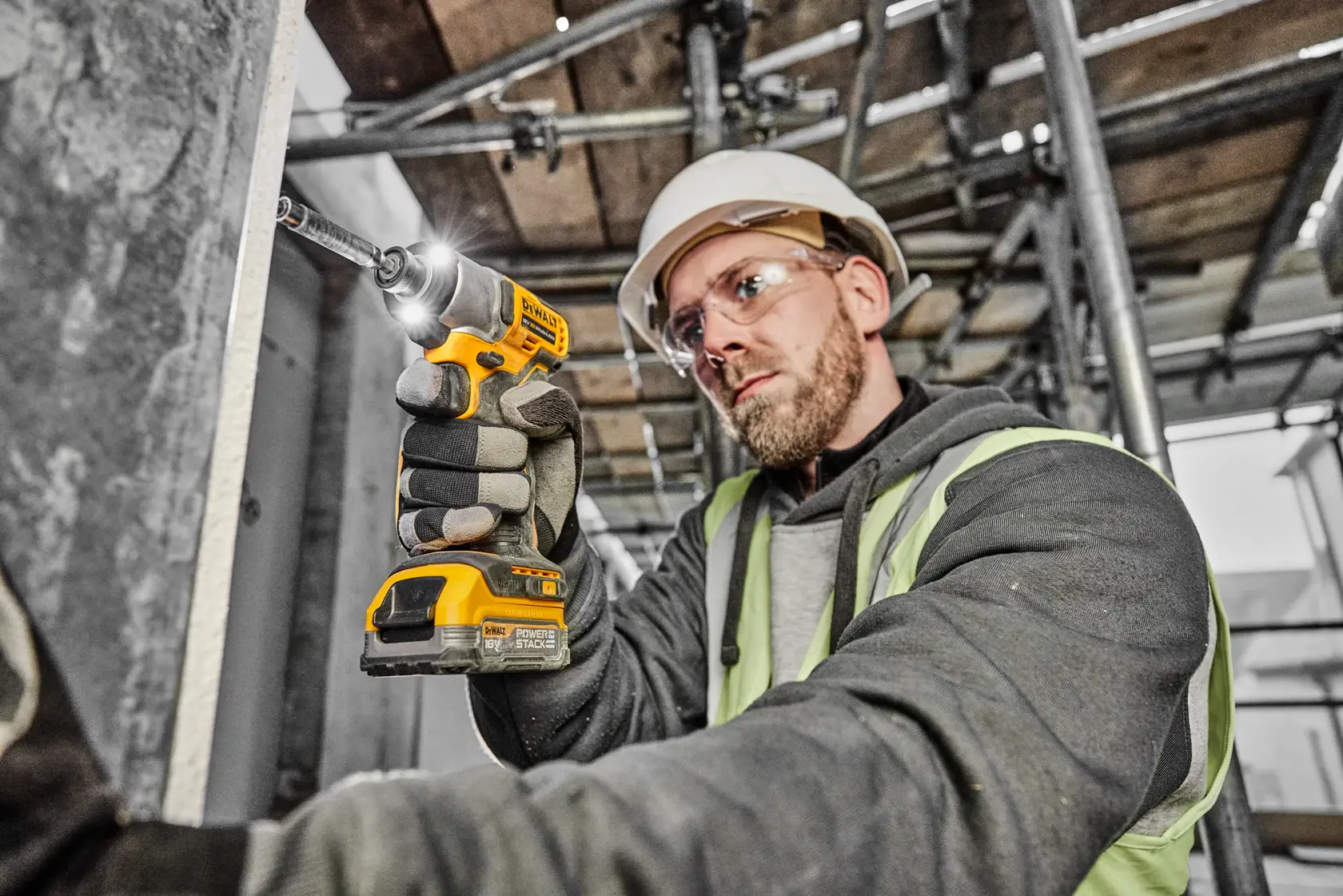 Eine Person mit Sicherheitsausrüstung verwendet einen gelben DEWALT Akkuschrauber, um eine Schraube in eine Metalloberfläche auf einer Baustelle zu drehen.