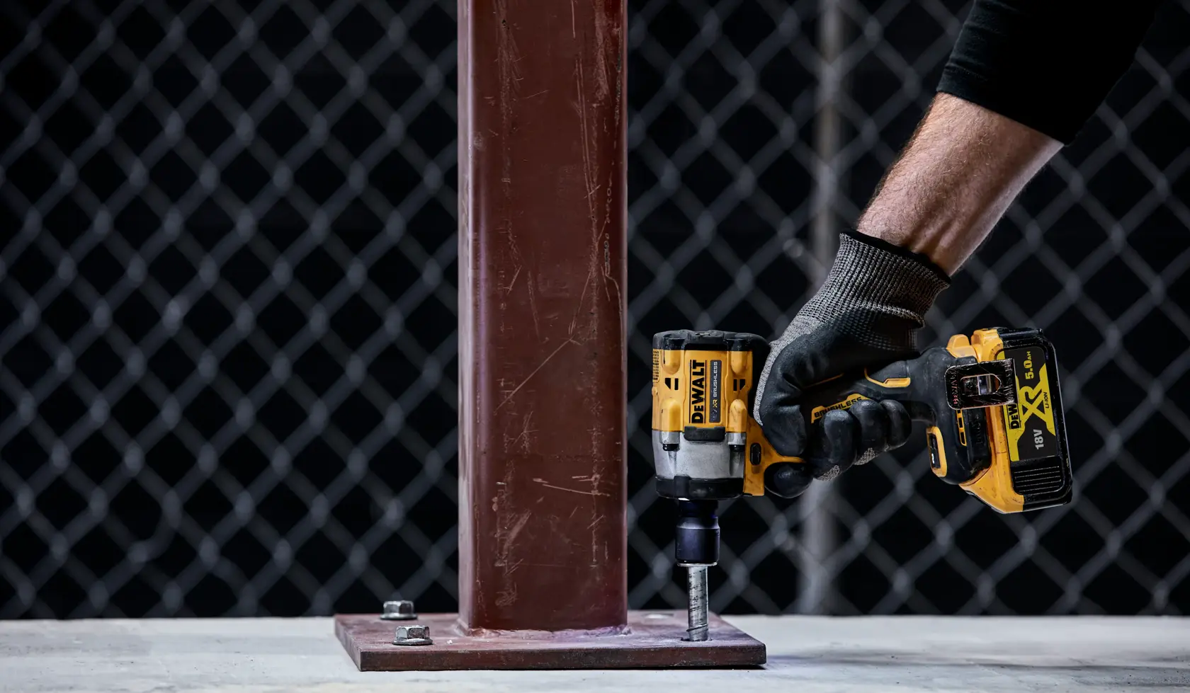Eine Person mit einem Arbeitshandschuh verwendet einen DEWALT DCF921NT Akku-Schlagschrauber, um eine Schraube am Fuß einer Metallstütze festzuziehen.
