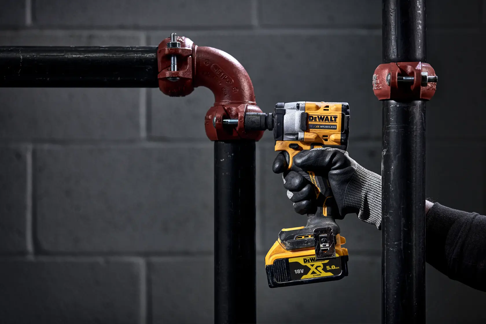 Eine Person mit einem schwarz-grauen Arbeitshandschuh verwendet einen DEWALT 18V Akkuschlagschrauber an einer schwarzen Metallrohrverbindung in einer industriellen Umgebung. Im Hintergrund ist eine graue Ziegelwand zu sehen.