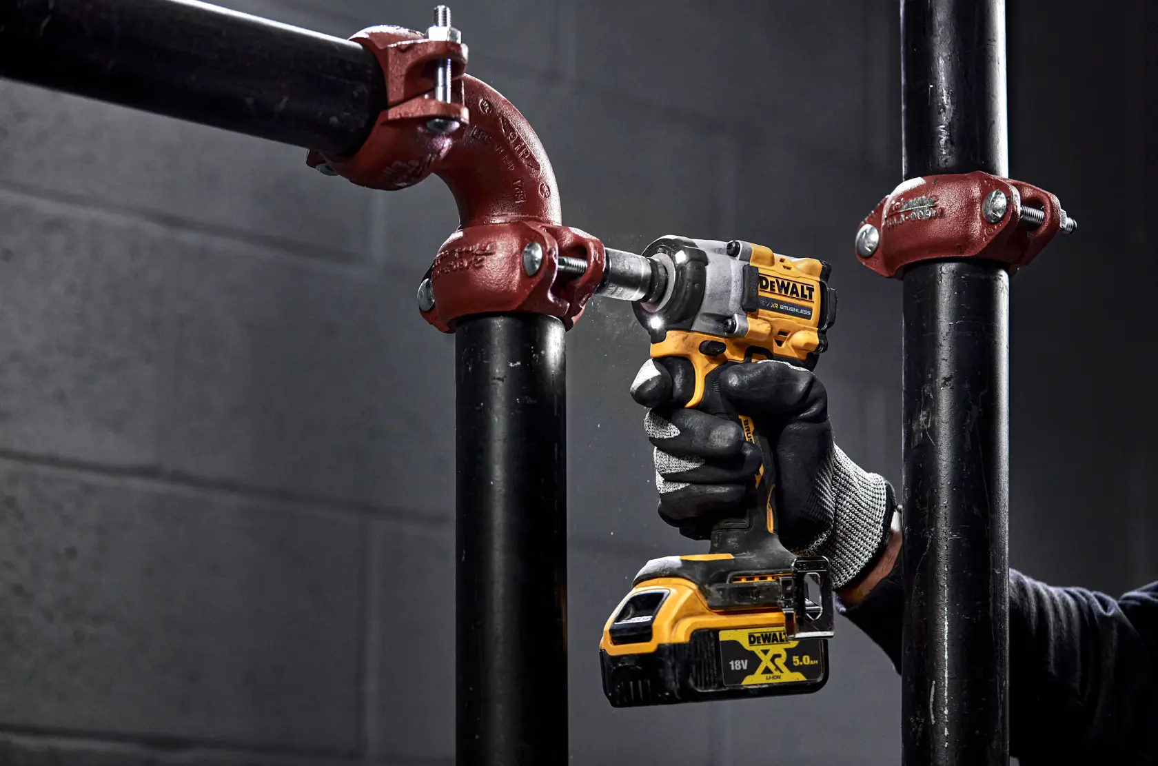 Eine Person mit Arbeitshandschuhen verwendet einen gelben DEWALT Akku-Schlagschrauber (Modell DCF922NT), um eine Metallrohrverbindung in einer industriellen Umgebung zu befestigen.