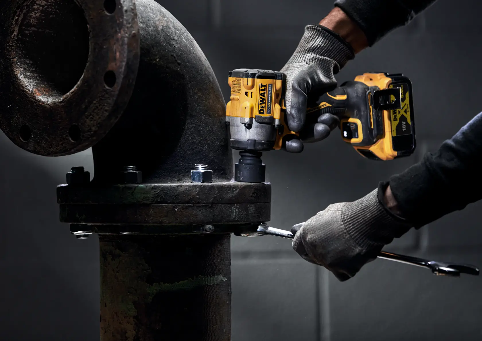 Eine Person mit Handschuhen benutzt einen kabellosen DEWALT Schlagschrauber, um Schrauben an einer großen industriellen Metallrohrverbindung festzuziehen.