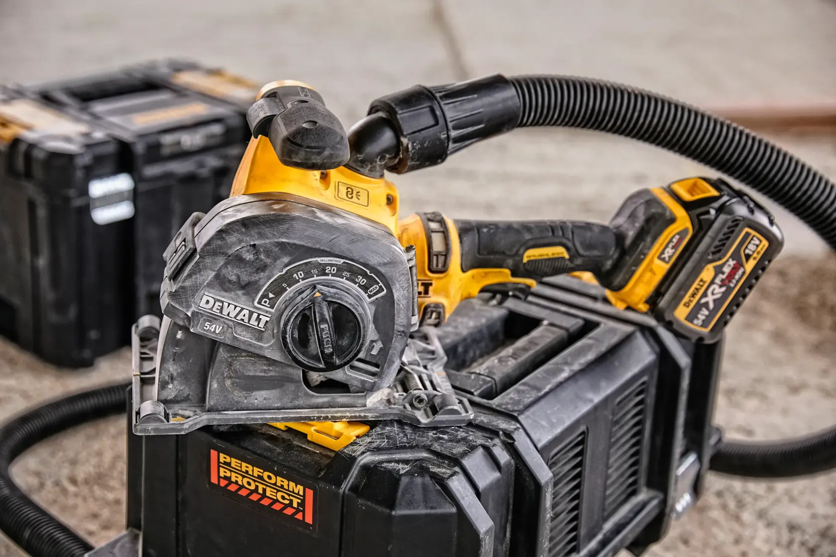 Imagen en primer plano de una herramienta eléctrica inalámbrica DEWALT, específicamente una herramienta de corte con una manguera de extracción de polvo conectada, situada sobre una caja de almacenamiento negra en un taller. Se puede ver otra caja de herramientas en el fondo.