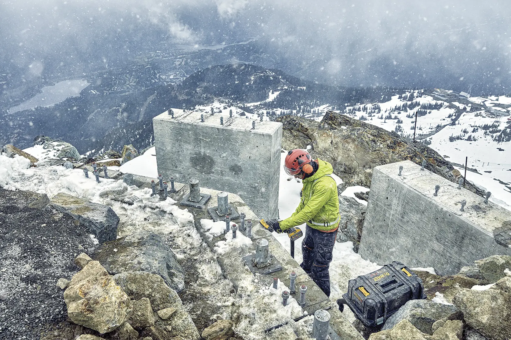 Trabajador usa herramienta DEWALT en obra de montaña nevada con bloques de concreto y maletín.