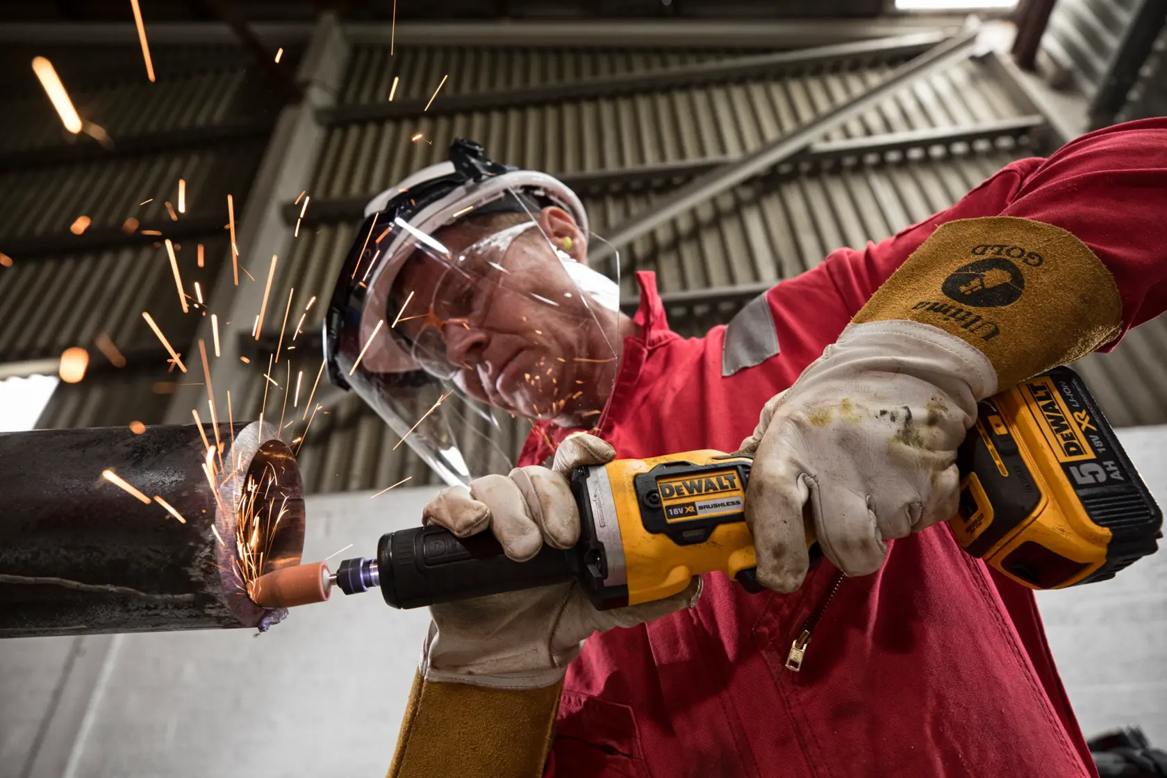 Una persona con guantes protectores y equipo de seguridad usa una amoladora inalámbrica DEWALT DCG426N sobre una tubería metálica, creando chispas en un entorno industrial.