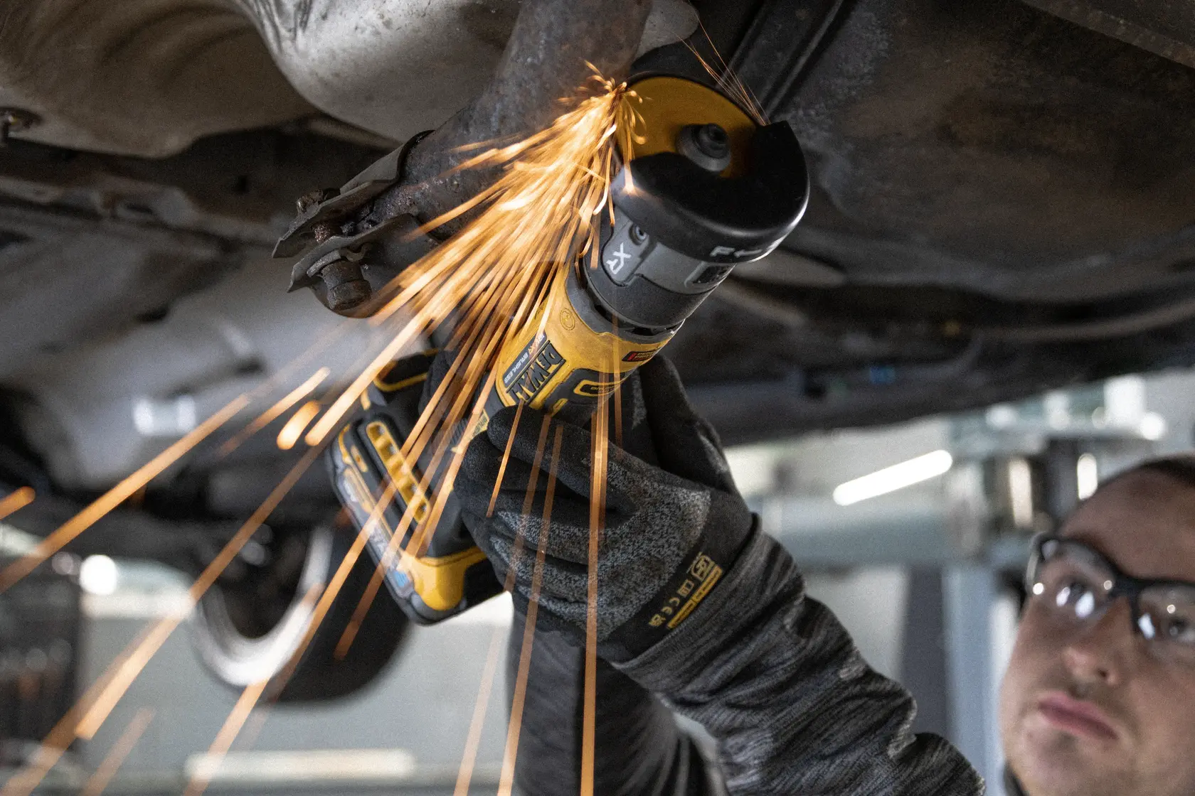 DEWALT DCG430N-XJ, DCG430N grinder in use under vehicle, sparks flying, worker hand with glove visible.