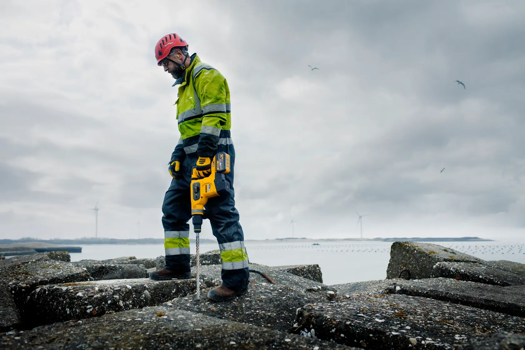 Una persona con chaqueta de alta visibilidad, casco de seguridad y guantes utiliza una herramienta eléctrica amarilla DEWALT sobre una superficie rocosa al aire libre. Se ven turbinas eólicas y agua al fondo bajo un cielo nublado.