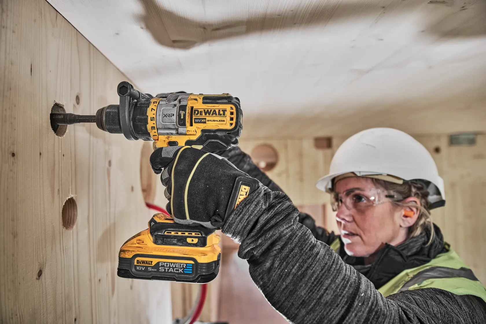 A person wearing a hard hat and gloves uses a DEWALT cordless drill to drill a hole in a wooden wall. The DEWALT Power Stack 18V battery is attached to the drill. The person's face is blurred for privacy.