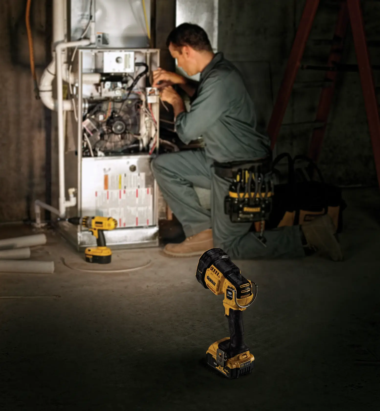 Un foco portátil inalámbrico DEWALT DCL043 está colocado en el suelo en primer plano, mientras un trabajador con mono realiza el mantenimiento de una unidad eléctrica en un entorno industrial con poca luz.