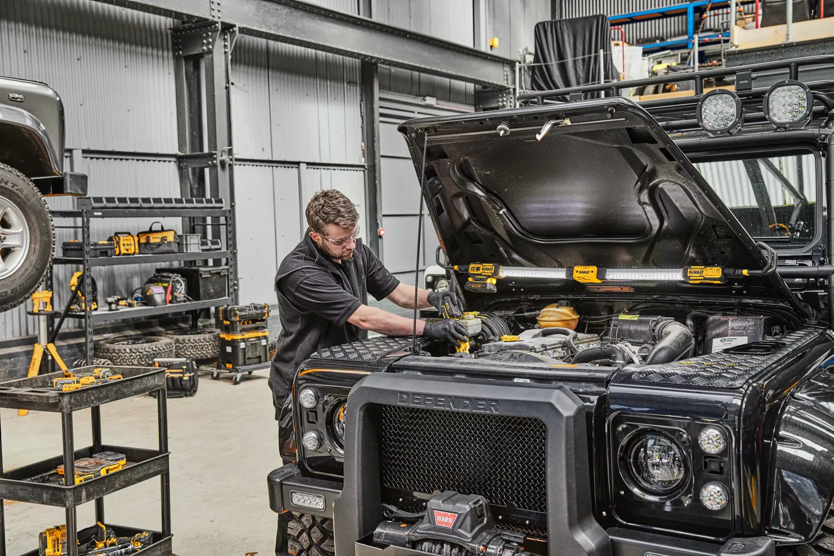 Una persona está trabajando en el motor de un Land Rover Defender negro dentro de un taller. Se pueden ver herramientas DEWALT en las estanterías y junto al vehículo, y la luz de trabajo DEWALT DCL045 está colocada bajo el capó abierto para iluminar el área del motor.