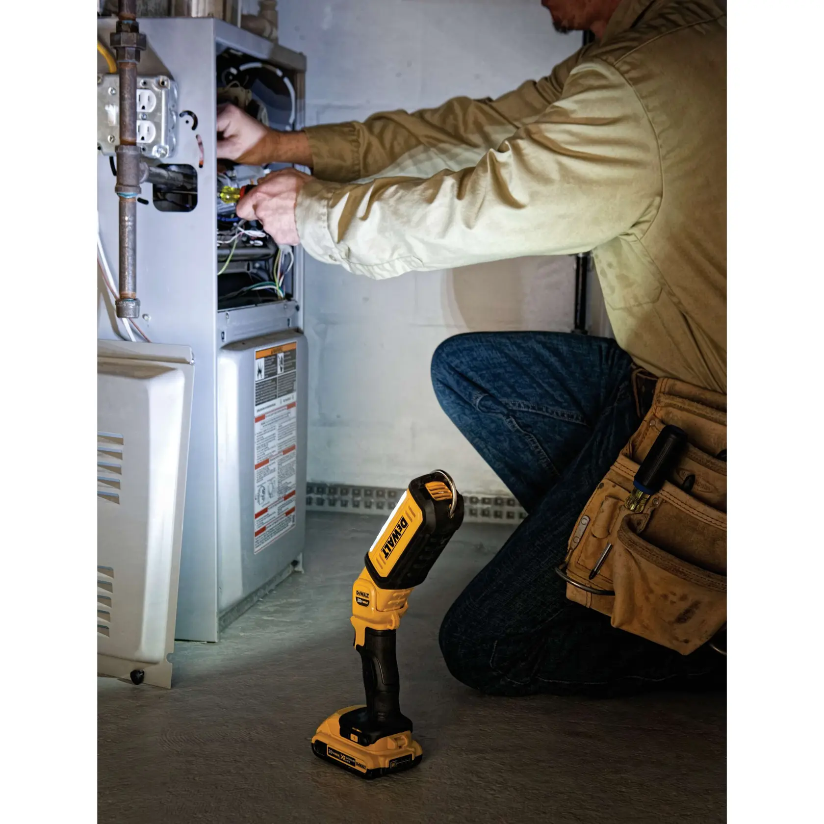 Eine Person kniet und arbeitet mit Werkzeugen an einer offenen elektrischen Einheit. Auf dem Boden steht eine tragbare DEWALT DCL050 LED-Arbeitsleuchte, die den Arbeitsbereich beleuchtet.