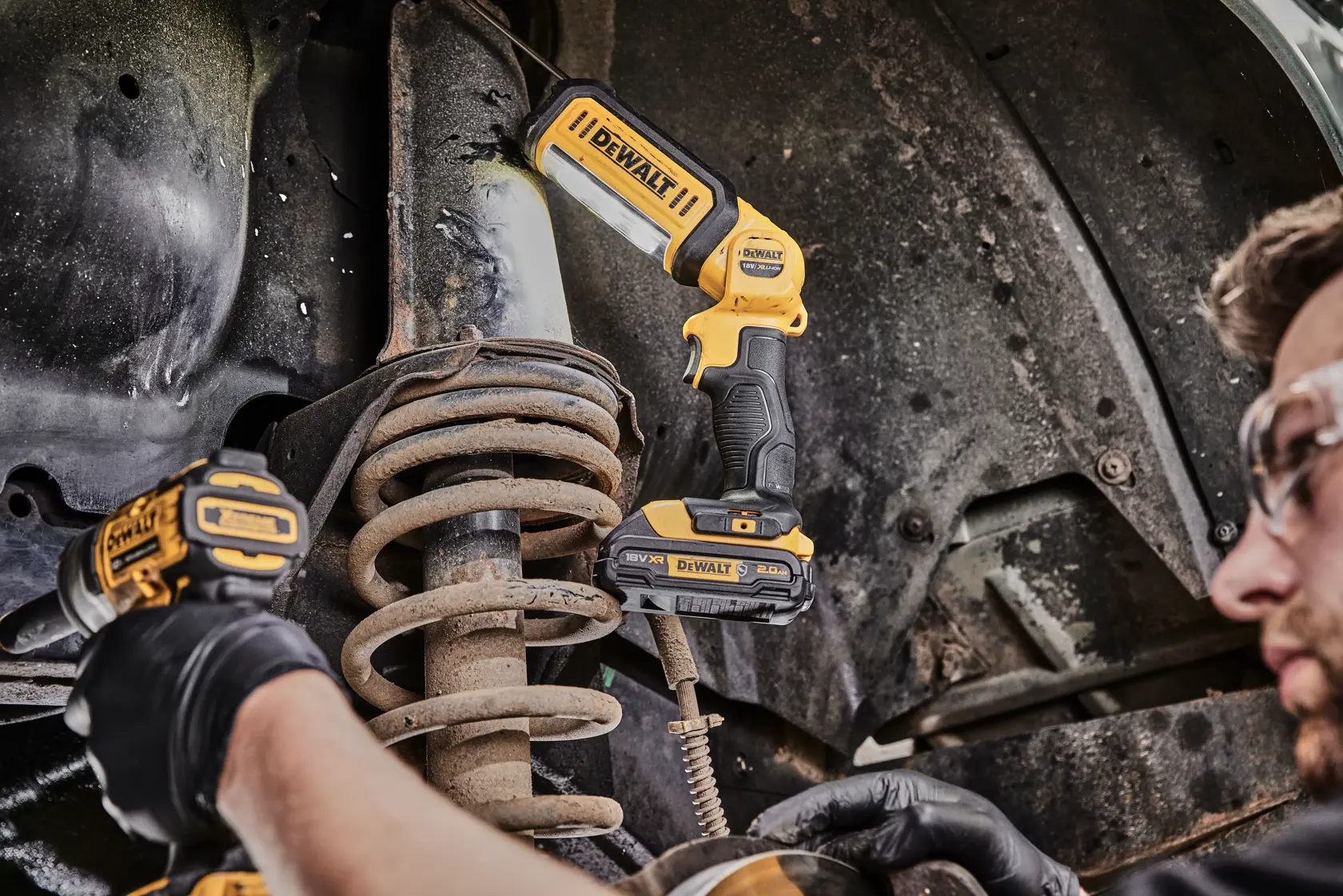 Eine DEWALT DCL050 LED-Arbeitslampe wird verwendet, um ein Automobil-Federungssystem zu beleuchten, während eine Person mit Sicherheitsbrille und Handschuhen mit einem weiteren DEWALT Werkzeug arbeitet.