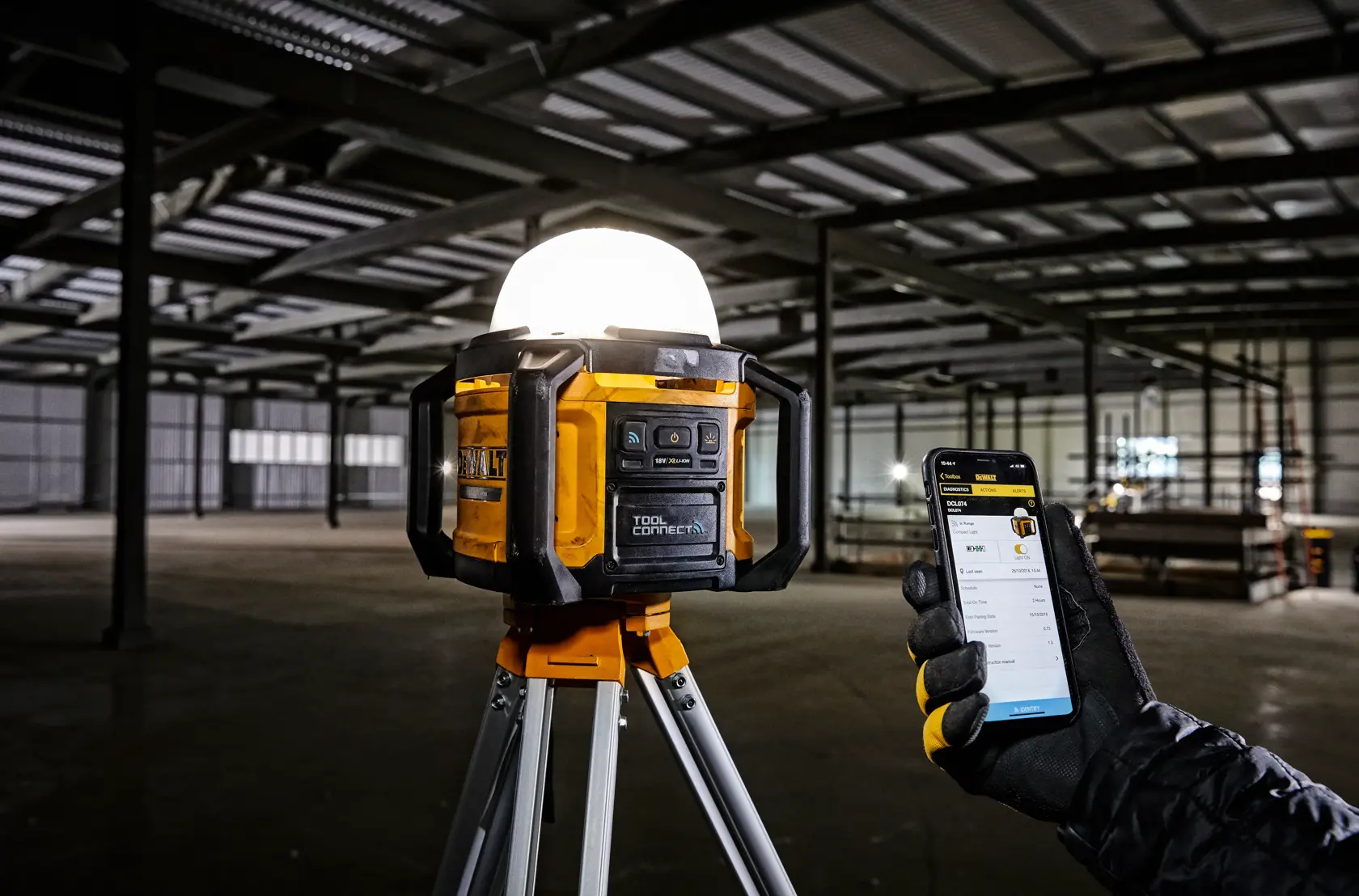 Luz de trabajo DEWALT en trípode en almacén, controlada por una mano con smartphone.