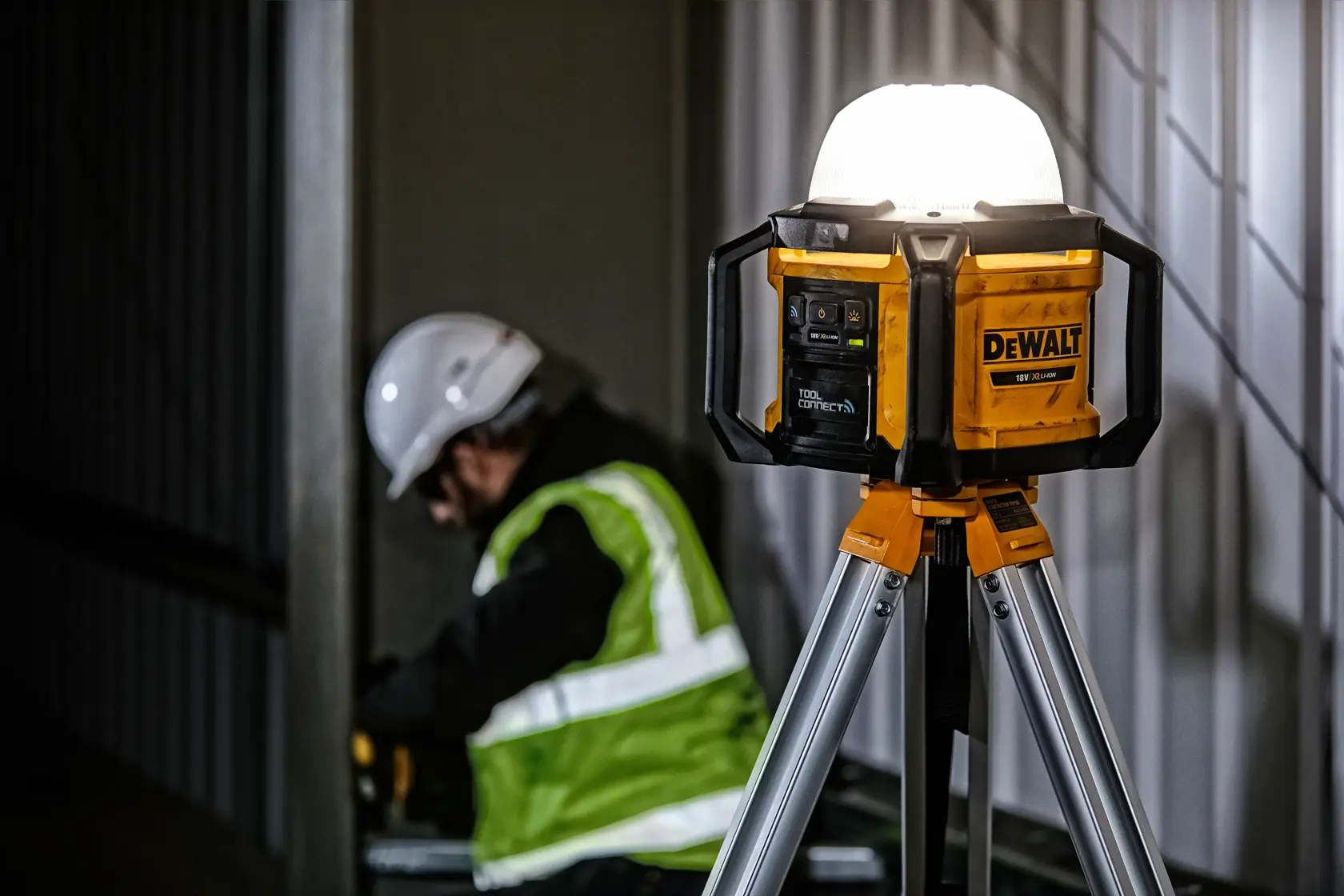 Luz LED DEWALT DCL074 en trípode en obra, trabajador con chaleco de seguridad visible al fondo.