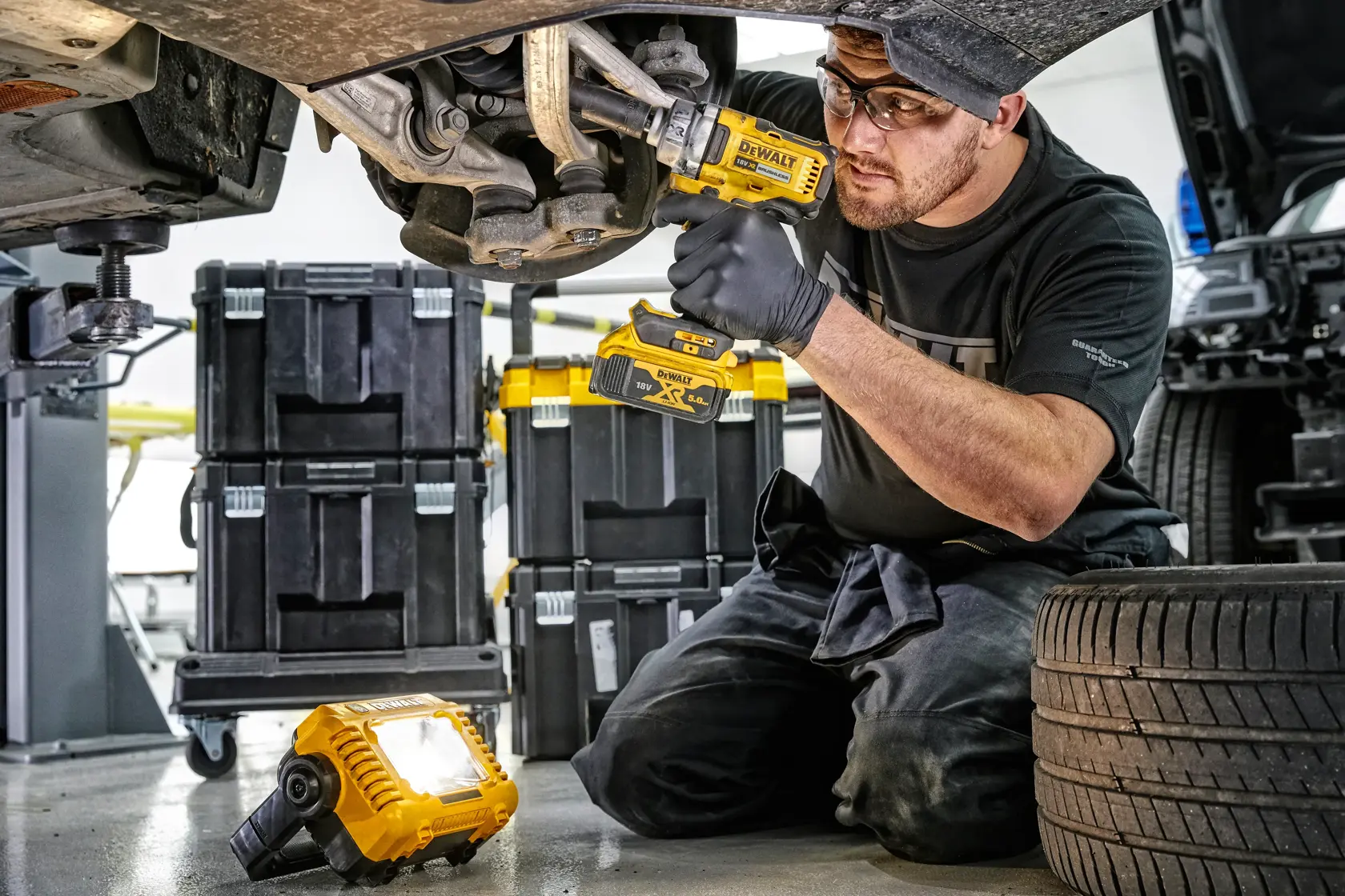 Mecánico usa herramientas DEWALT y luz LED bajo un auto, cajas de herramientas visibles atrás.