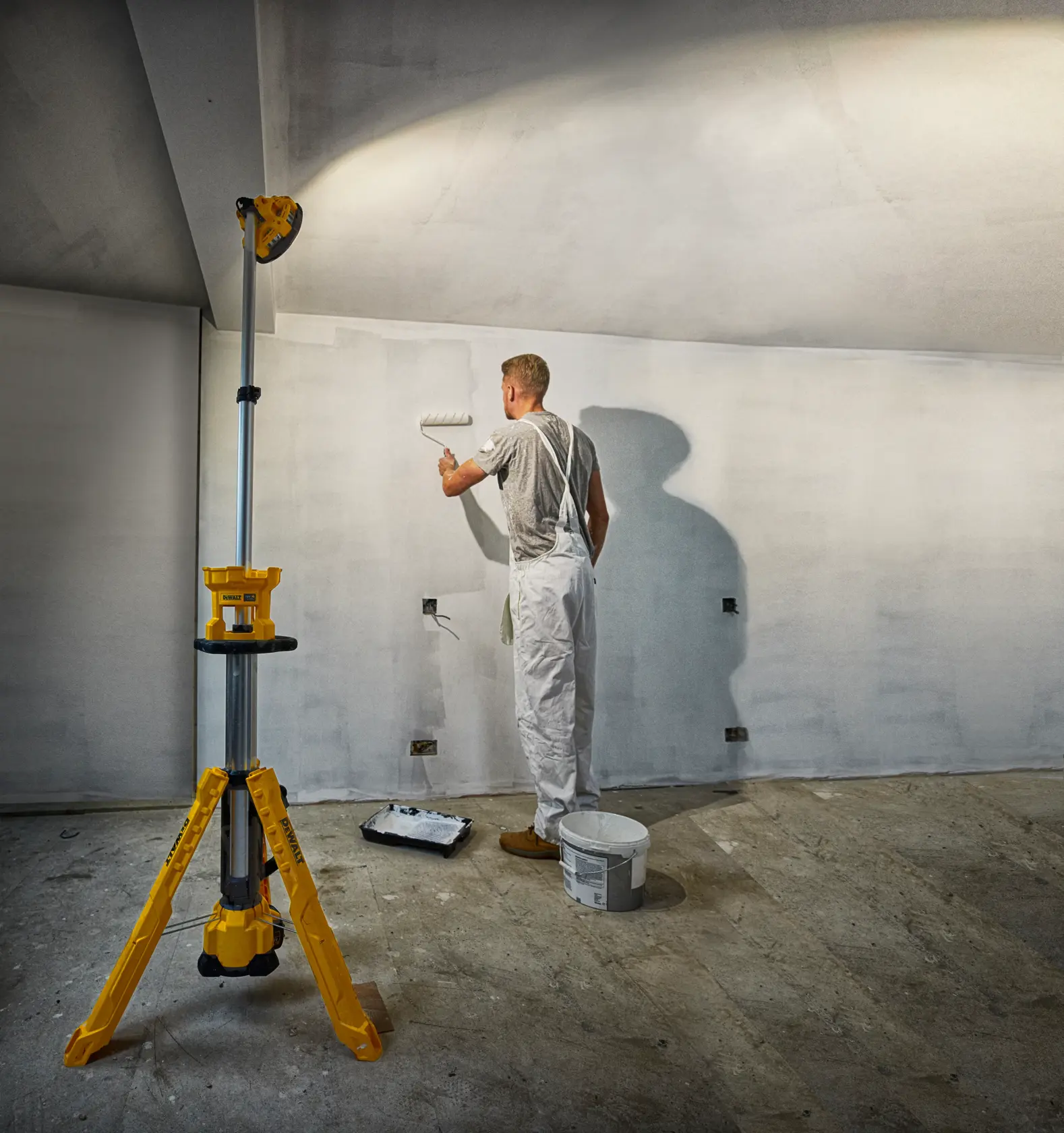 A painter uses a paint roller on a wall, illuminated by a yellow DEWALT tripod work light in a room under renovation.