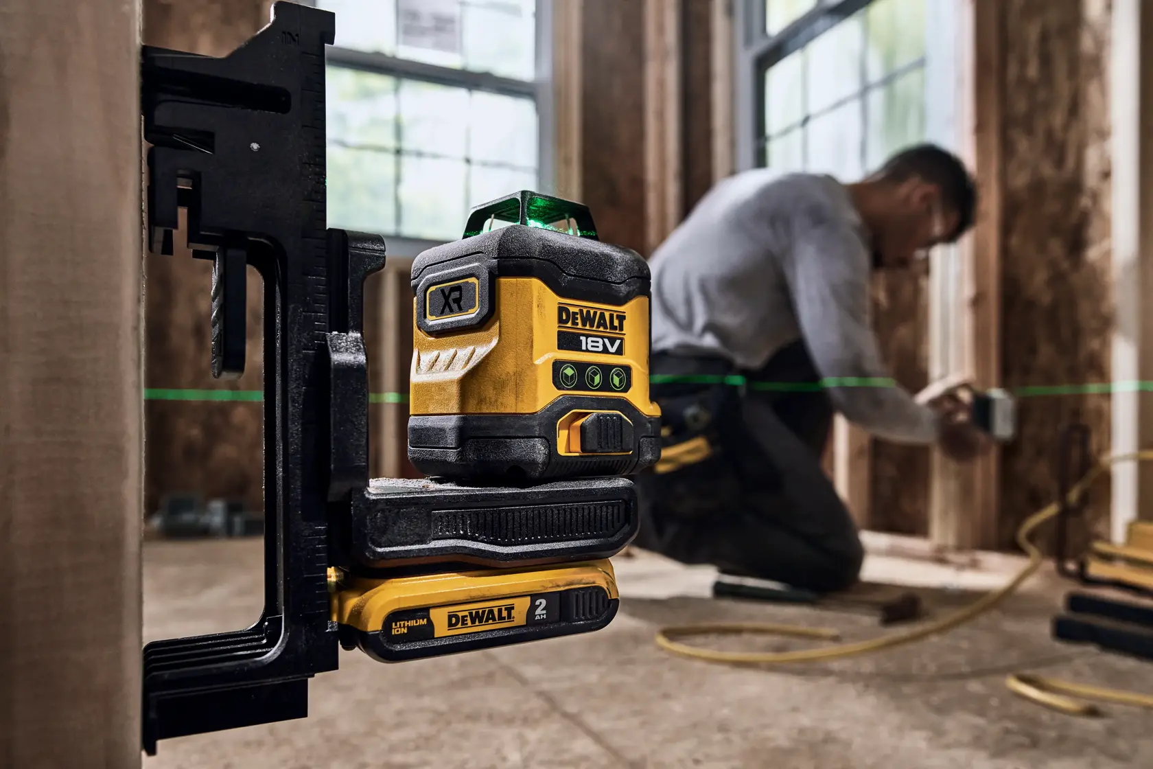 Nivel láser de línea verde DEWALT 18V XR fijado en un soporte con batería, usado en una obra de construcción. Una persona está trabajando en el fondo con el rostro desenfocado y la línea láser verde es visible en el espacio de trabajo.