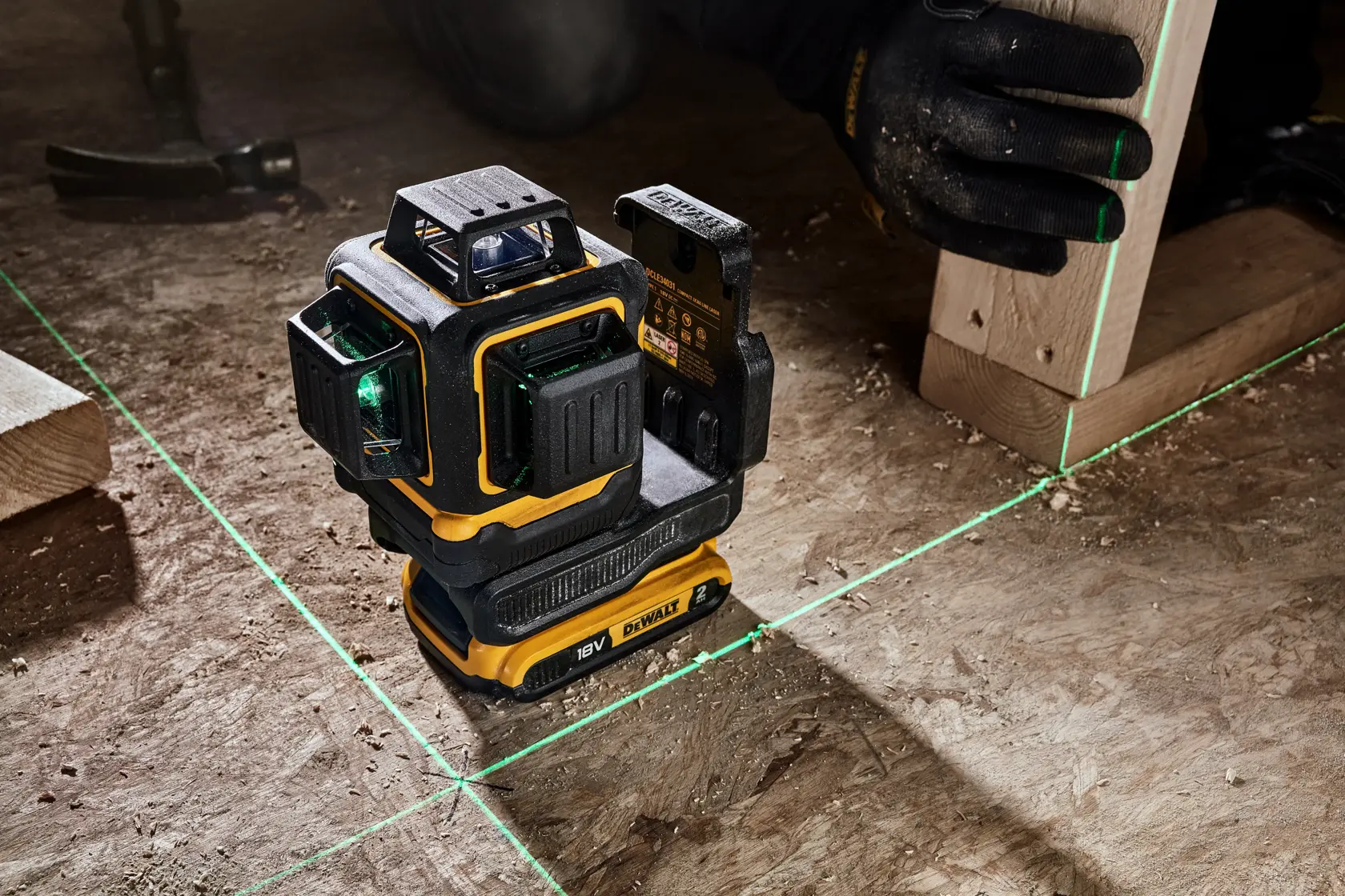 Un nivel láser DEWALT DCLE34031N 18V proyecta líneas láser verdes sobre una superficie de construcción. Una mano enguantada sujeta una viga de madera, alineándola con las líneas láser. Herramientas y materiales de construcción son visibles al fondo.