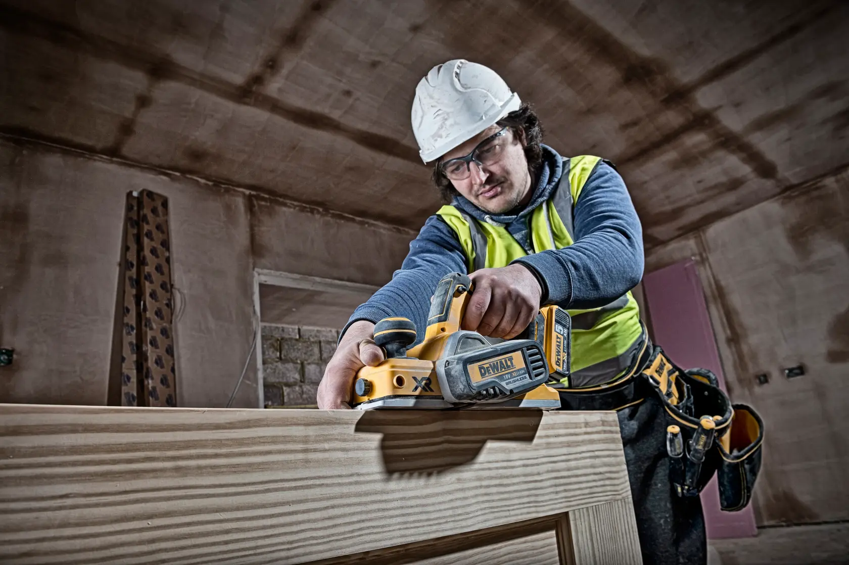 Photo of worker using DEWALT cordless planer DCP580N on wood indoors, wearing safety gear.