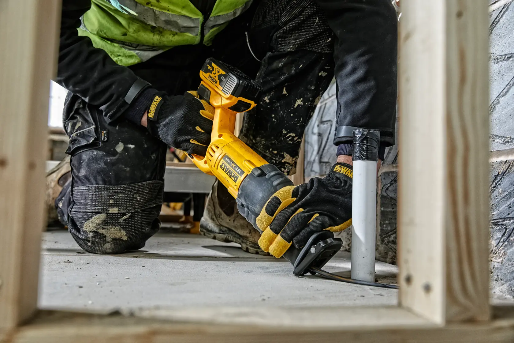 Worker using yellow DEWALT cordless reciprocating saw to cut metal pipe on construction site floor.